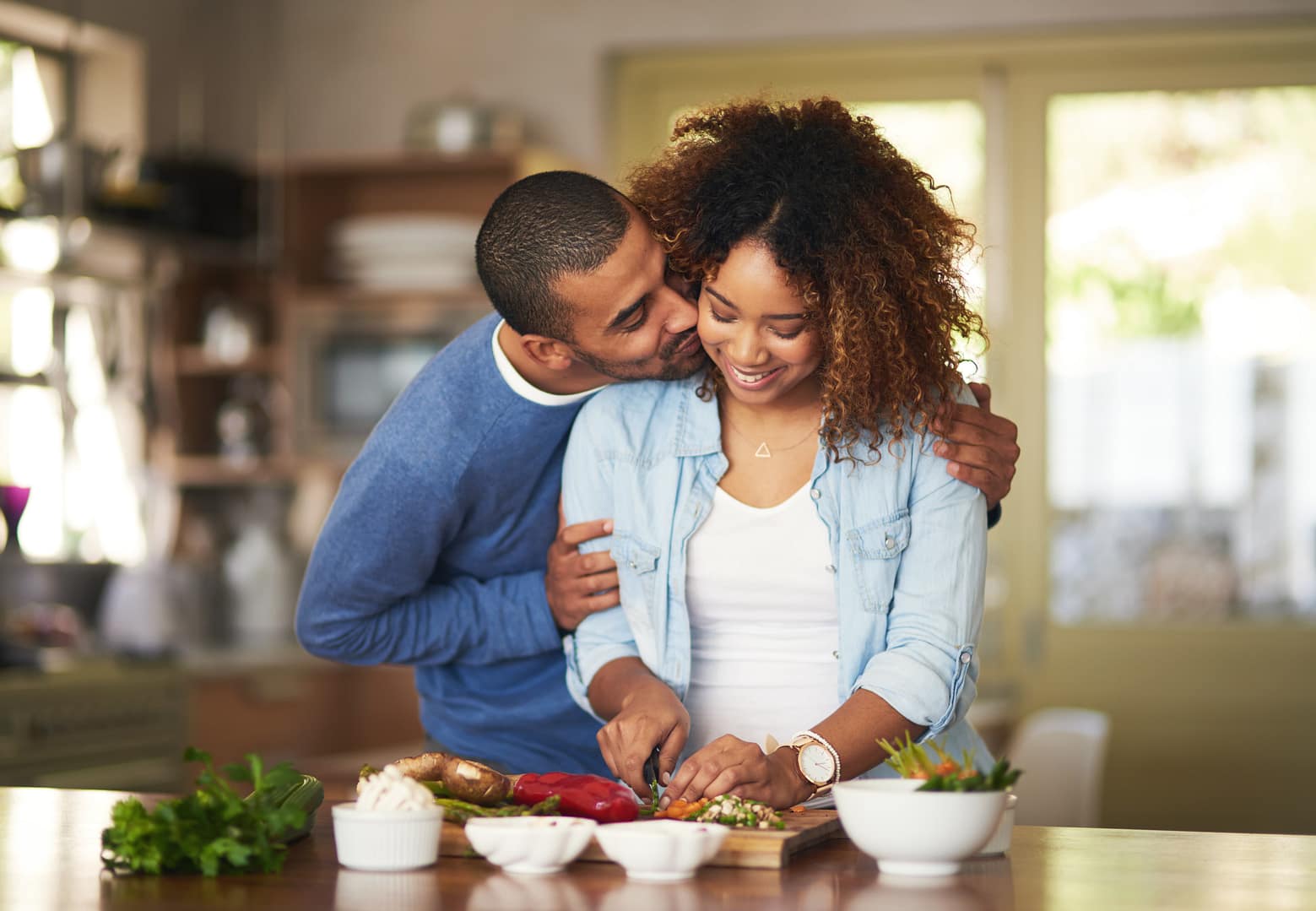A man hugs a woman from behind as she chops vegetables on a kitchen counter, both smiling and enjoying quality time in marriage. Fresh ingredients and bowls are arranged on the counter.