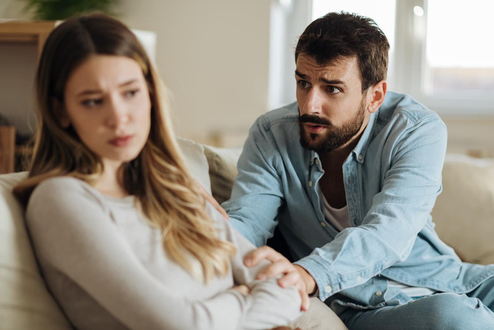 Man holding his wife’s hands while apologizing during an emotional conversation on the couch, representing a couple working through conflict with relationship repair skills.