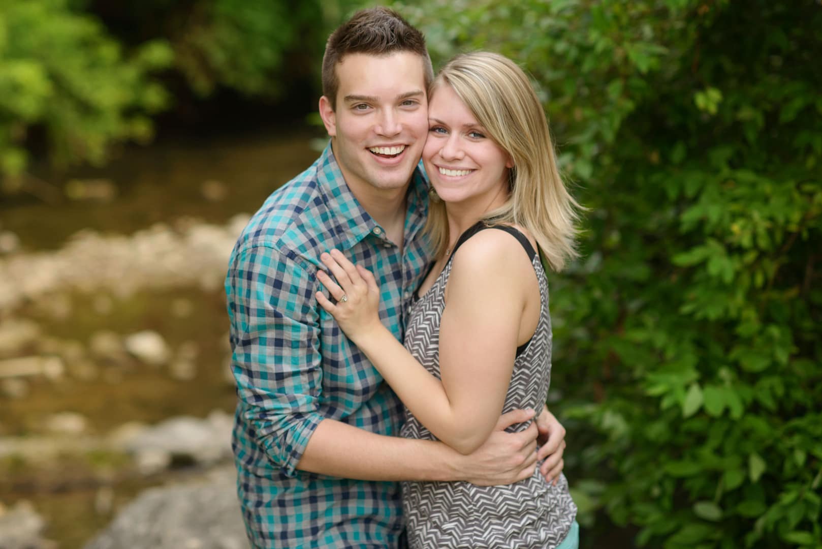 A smiling couple stands close together outdoors, surrounded by greenery, a stream, and rocks—capturing a moment of apology and forgiveness in marriage.
