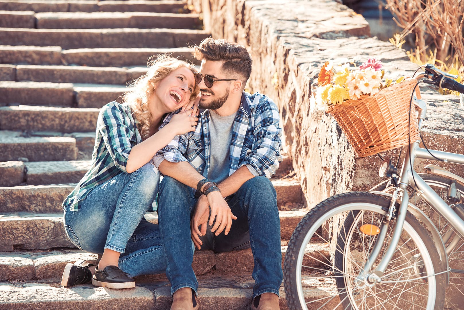 A couple sits on outdoor stone steps, smiling and laughing together, embodying the joy of intentional dating. Nearby, a bicycle with a basket of flowers adds to their charming moment.