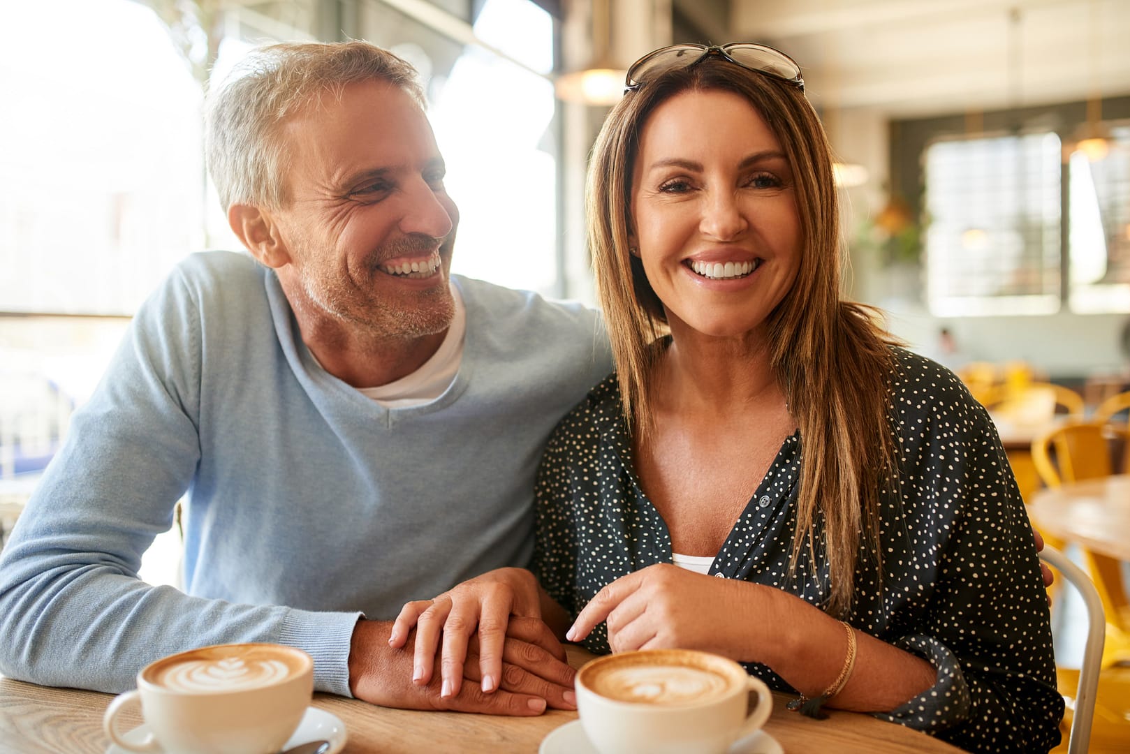 A man and woman sit together at a table in a café, smiling and holding hands over two cups of coffee—a perfect scene for those seeking micromance ideas for men.