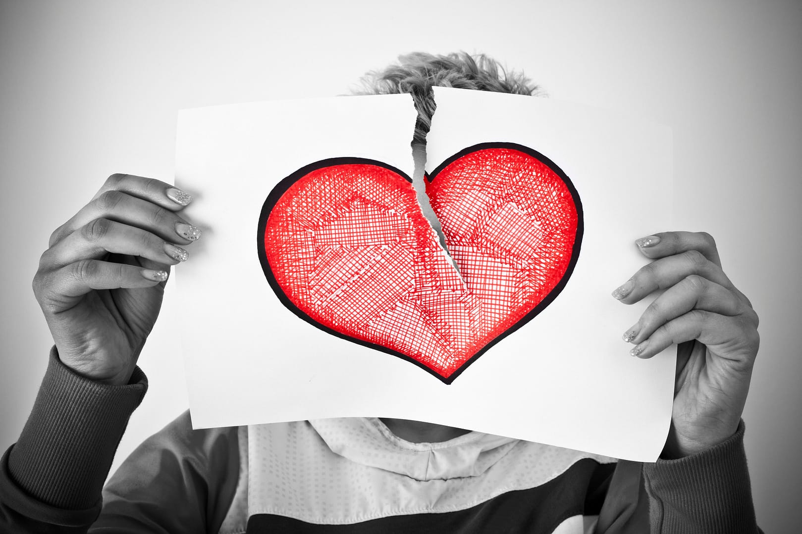 A person holds up a piece of paper with a red heart drawn on it, torn in half, obscuring their face from view, capturing the emotions of What to Do After a Breakup.