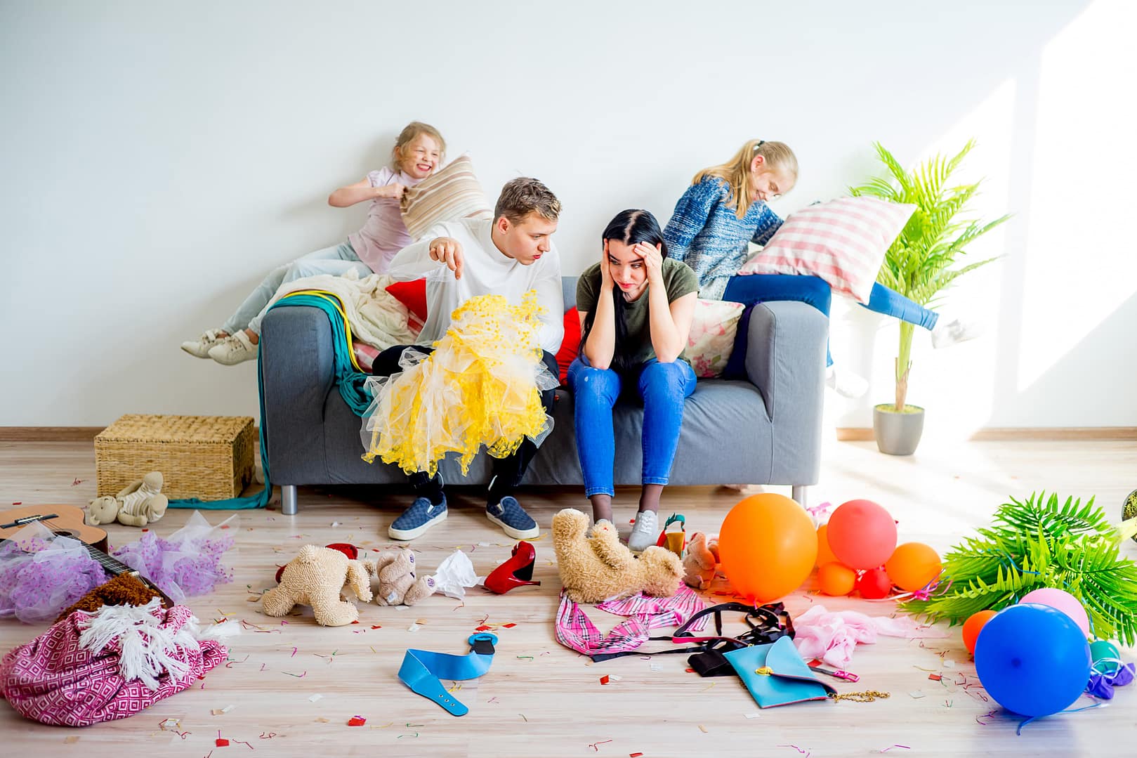 Two adults sit with their heads in their hands on a messy couch, worn out from managing the division of labor in marriage, while two children play energetically amid toys, balloons, and scattered items in a cluttered living room.