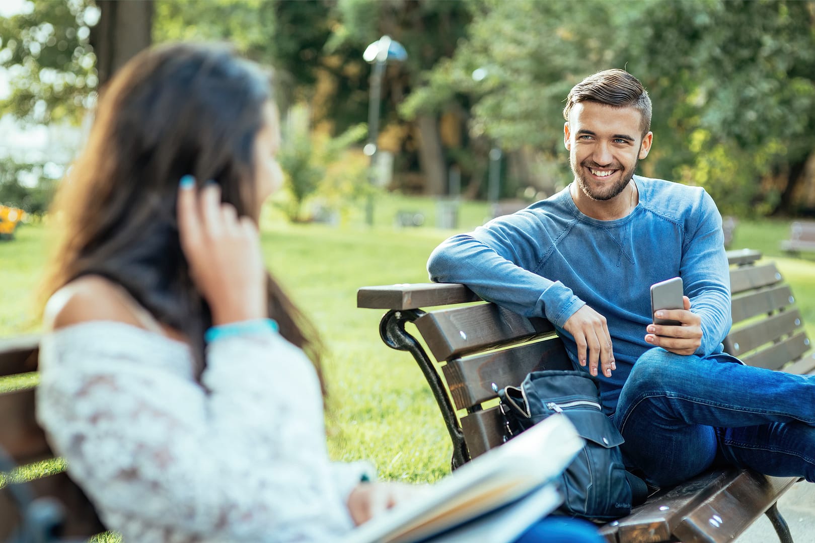 Man looking at a woman at the park. They both sitting in a different park bench but next to each other. They both are being flirty to each other.