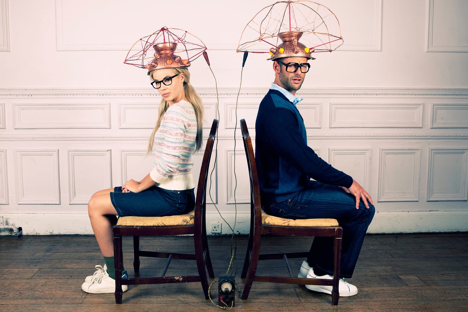 A woman and a man sit back to back on chairs, wearing wired metal helmets connected by cables, perhaps exploring unique Micromance ideas for men or deepening their understanding of men in a room with white walls and wooden flooring.