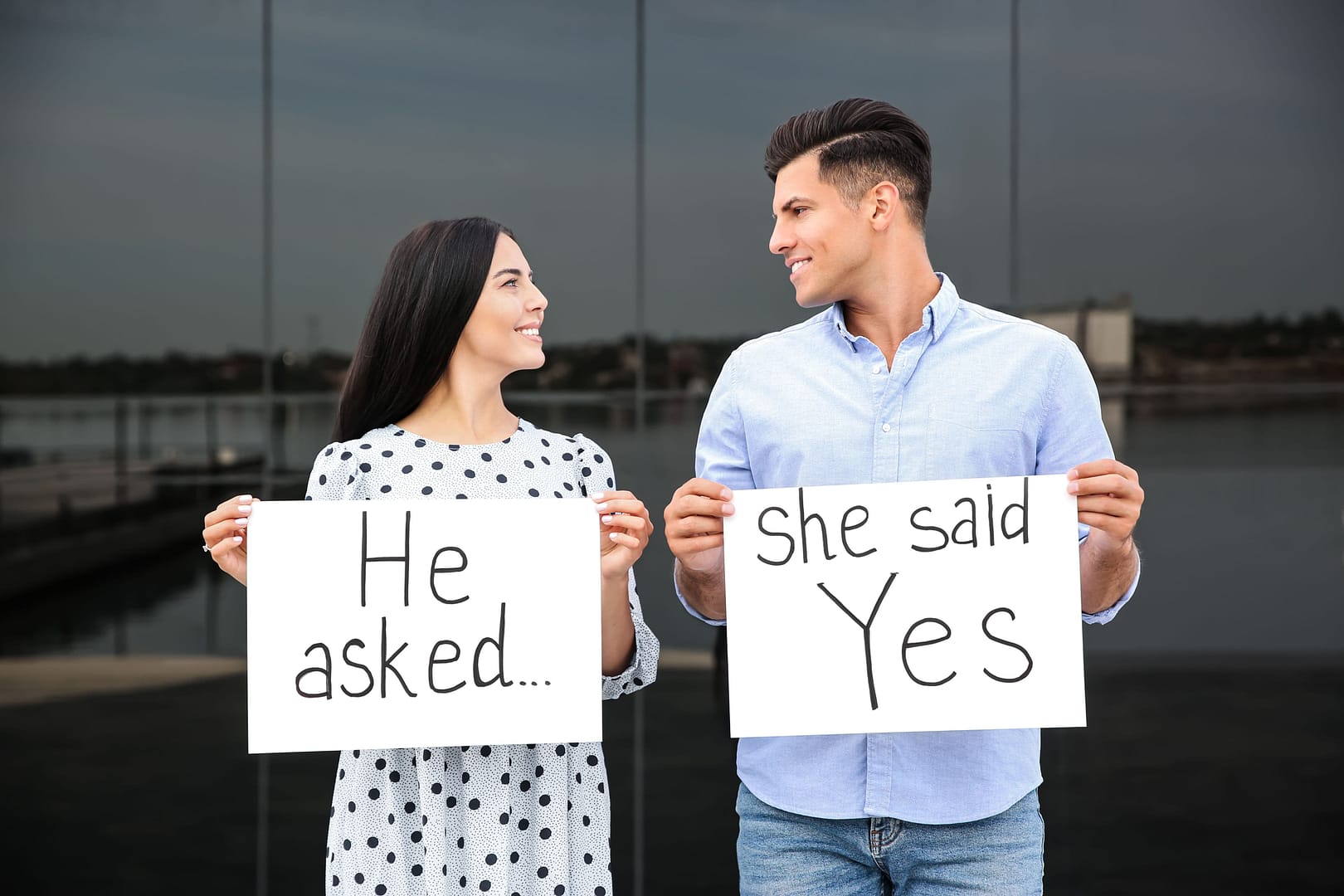 Man and woman smiling at each other while holding engagement announcement signs - questions to ask before marriage
