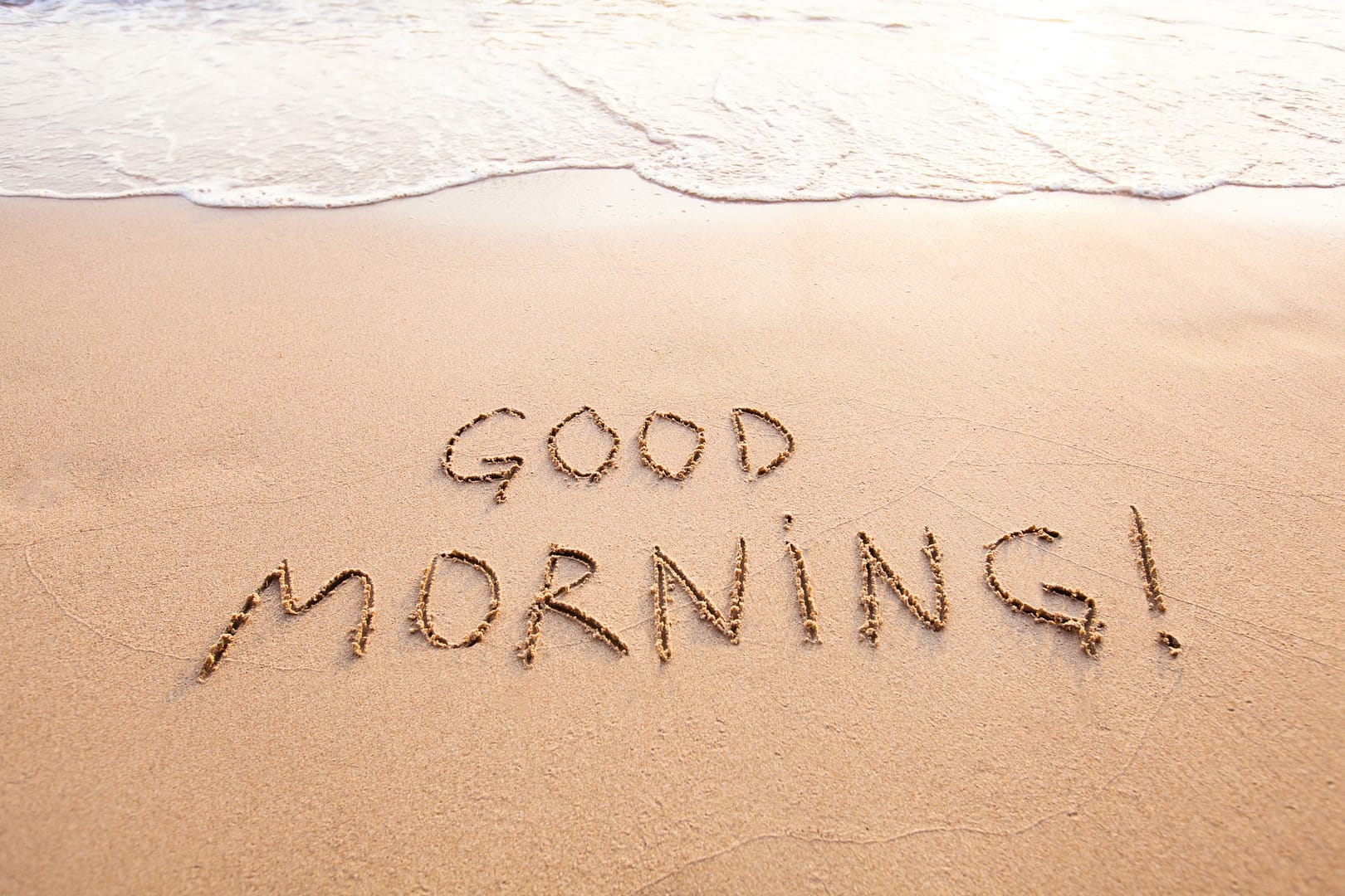 The words "GOOD MORNING!" are written in the sand near the edge of the ocean, with gentle waves approaching the shore. Flirty good morning messages for him
