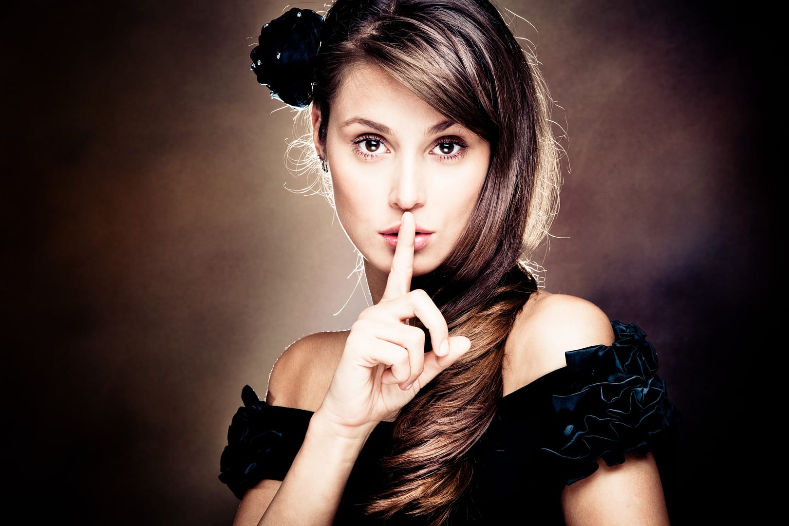 A woman with long brown hair makes a "shh" gesture, embodying the intrigue of His Secret Obsession, as she poses in a black off-shoulder dress with a flower hair accessory against a dark background.
