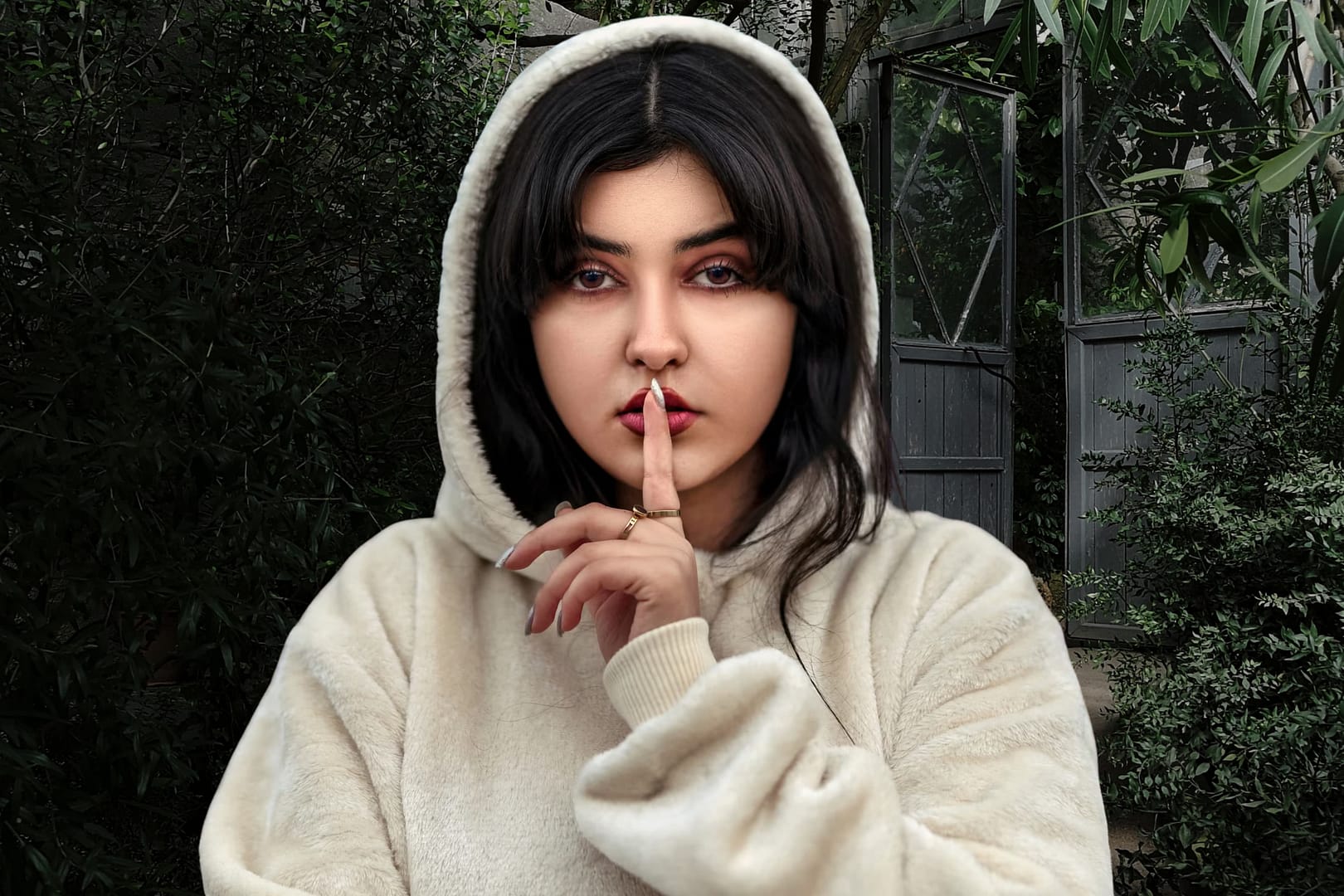 A person in a beige hoodie stands outdoors, holding a finger to their lips in a gesture signaling silence—capturing the mysterious allure of Secret Obsessions Men—amid greenery and a glass door in the background.