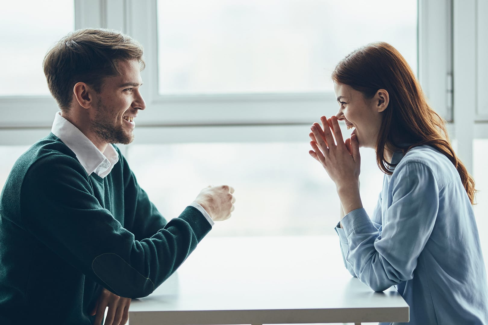A man and a woman sit across from each other at a table, smiling and laughing in a bright room with large windows, showing how communication tips for couples can strengthen their connection.
