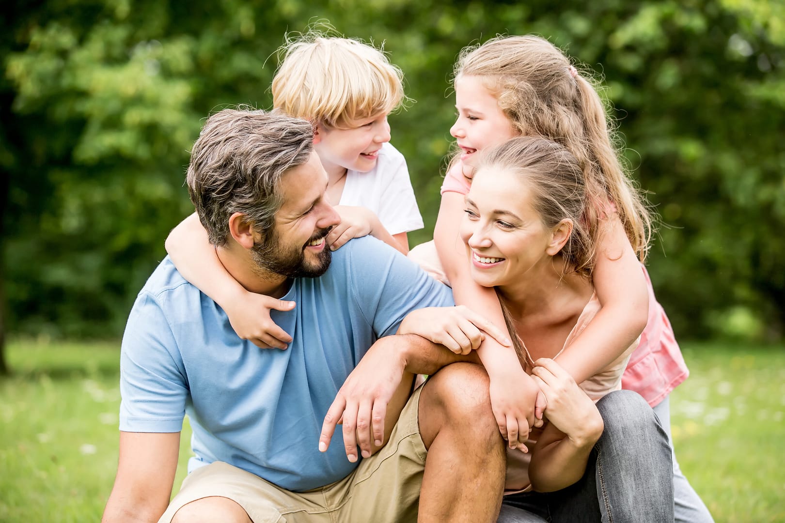 A man and woman sit outdoors while a boy and girl hug them from behind, all smiling, with green trees in the background—capturing a joyful moment for families focused on long-term goals for married couples.