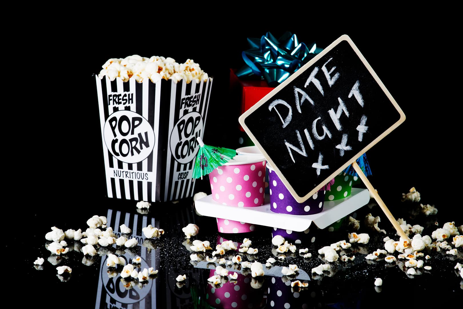 Popcorn in a striped container, colorful gift boxes, and a chalkboard sign reading "Date Night" on a black background, with scattered popcorn around—setting the perfect mood for a cozy evening of soft dating.