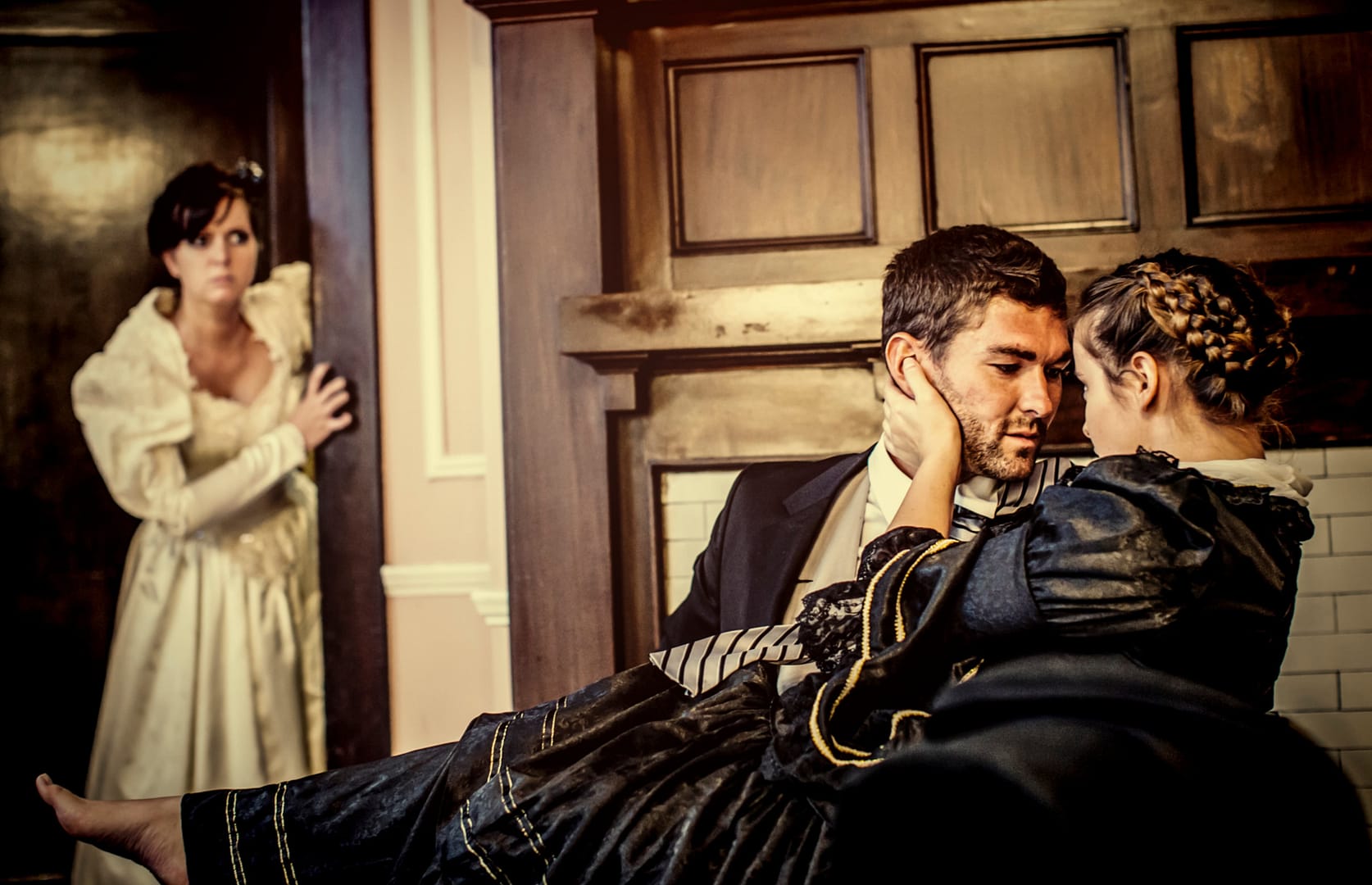 A woman in a white dress watches from a doorway as a man and a woman in period costumes share an intimate moment by a fireplace. Marriage after betrayal