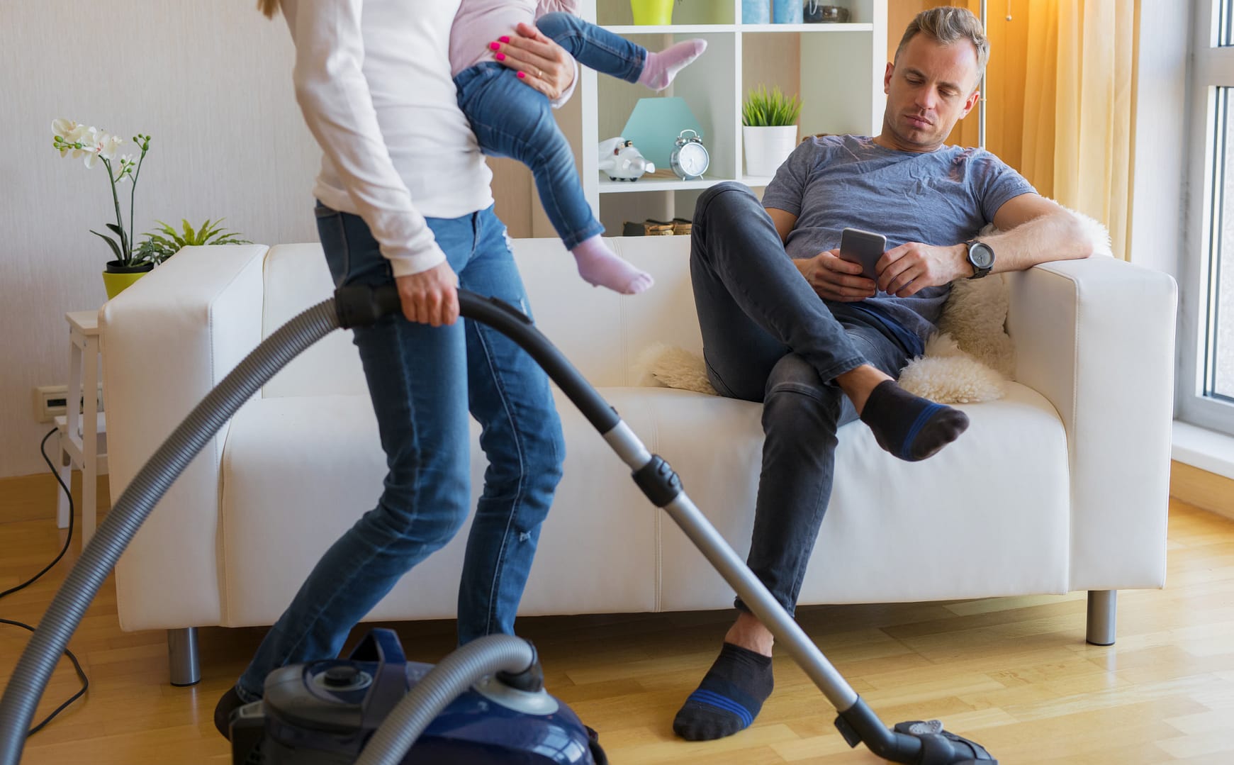 A woman holding a child vacuums the floor while a man sits on a couch using his smartphone, subtly illustrating the hidden cost of household inequality in the division of labor within marriage.