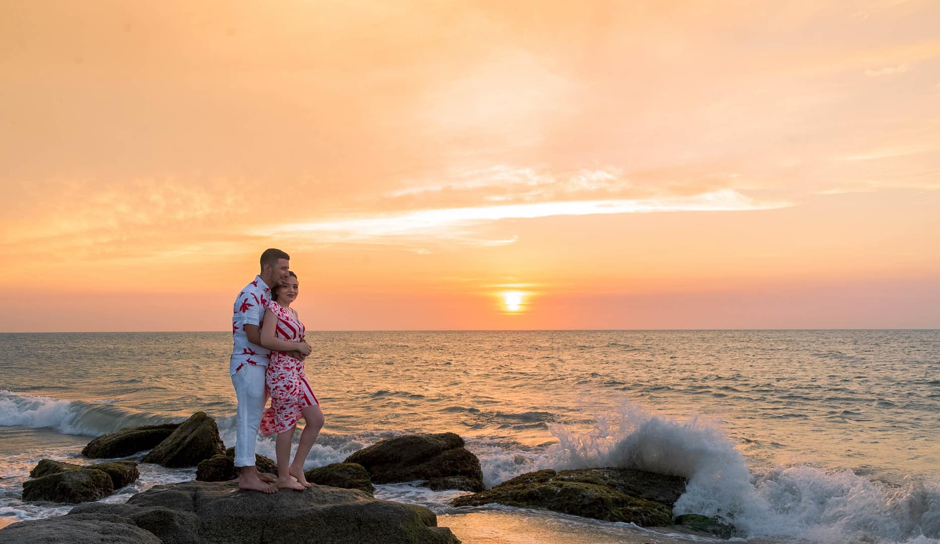 A couple stands on rocks by the ocean, embracing with patience in relationships as waves crash nearby and the sun sets, painting an orange sky in the background.