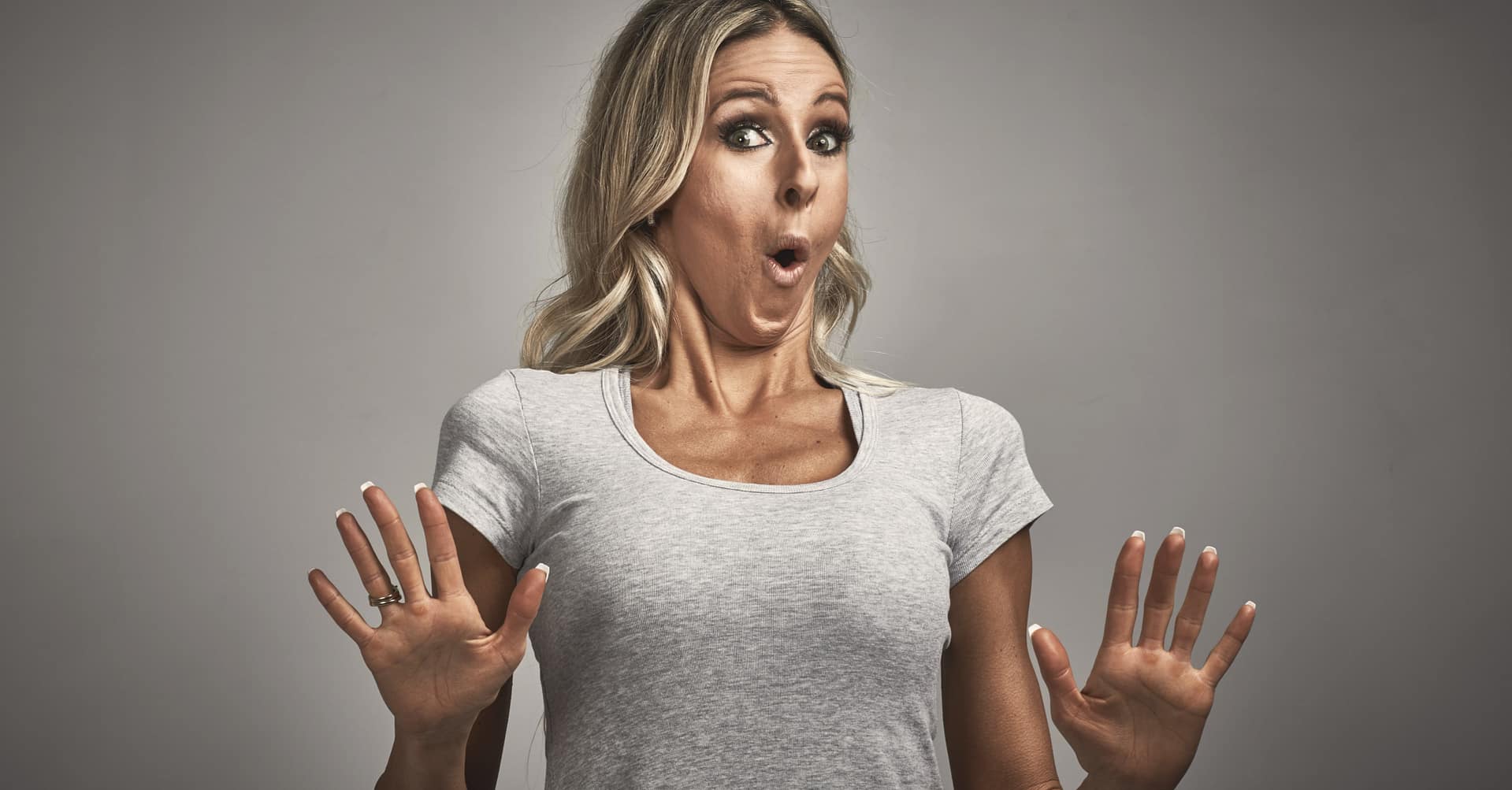 A woman with long blonde hair wearing a grey t-shirt appears surprised, with wide eyes and mouth open, holding her hands up in front of her chest—capturing the shock of realizing key Relationship Mistakes to Avoid.