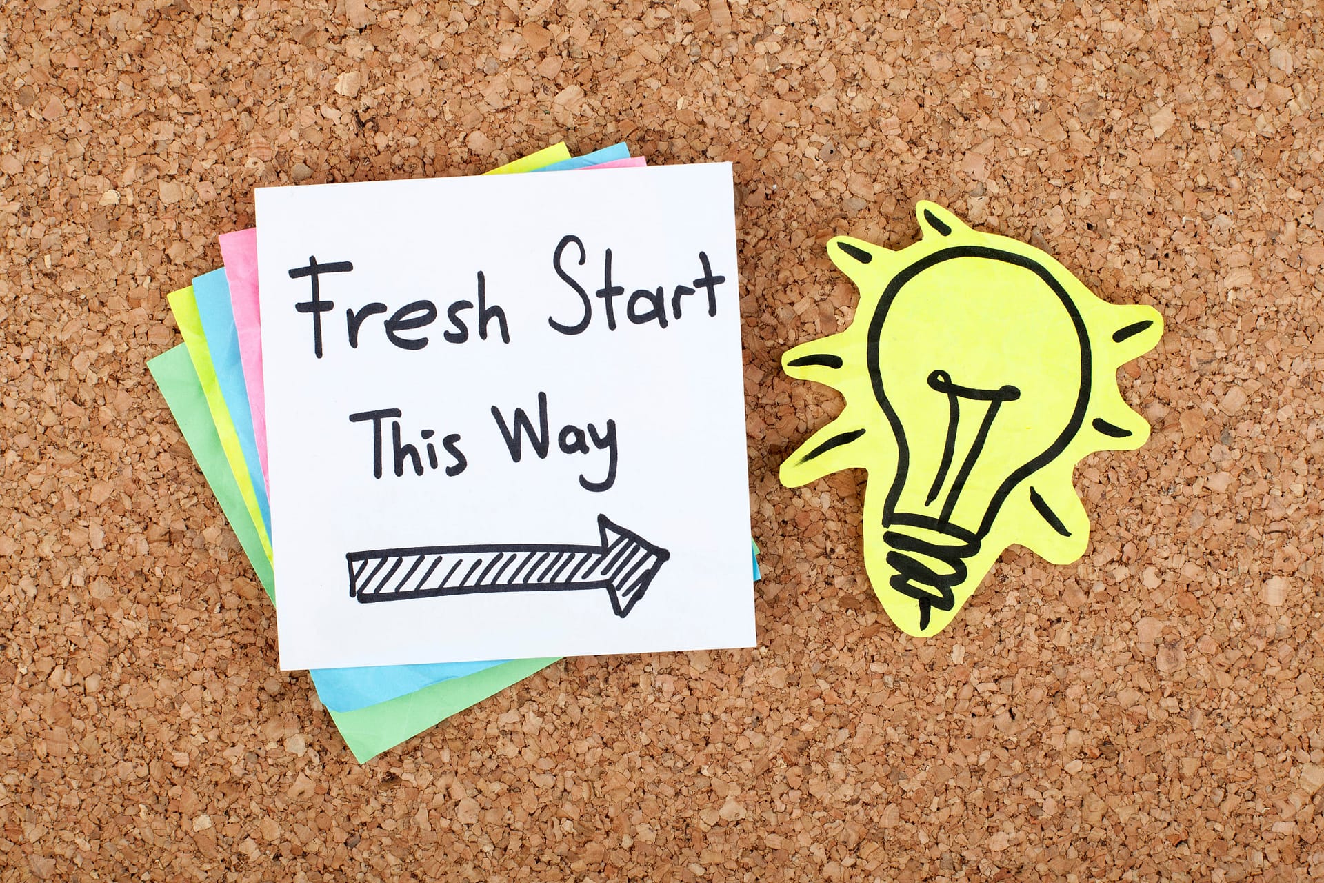 A stack of sticky notes on a corkboard, with the top note reading "fresh start this way" and an arrow, perfect for quotes for women embracing change. Next to it sits a yellow paper cutout of a lightbulb. New beginnings and fresh starts