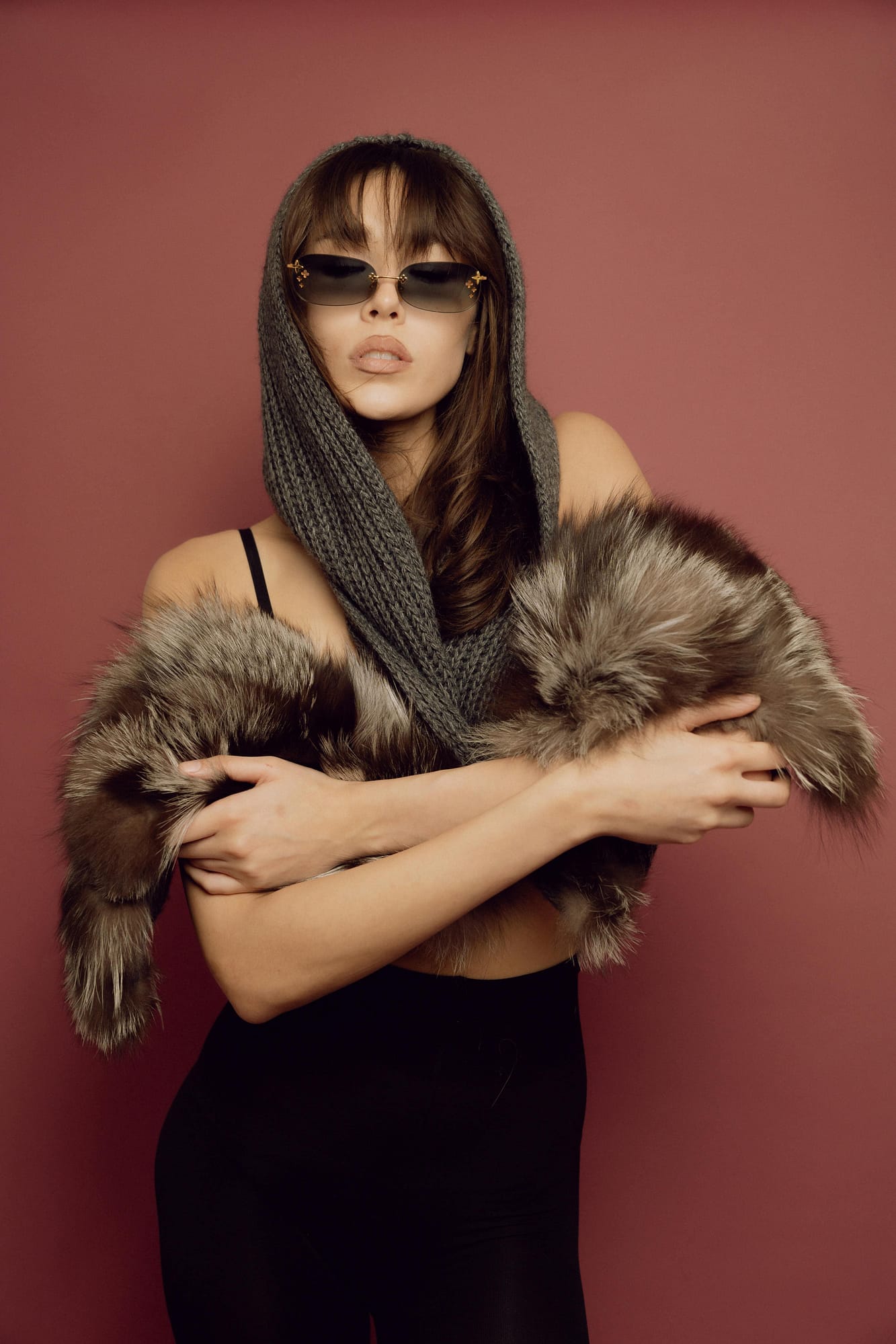 Woman wearing sunglasses and a knitted hood poses against a reddish background, draped in a faux fur garment