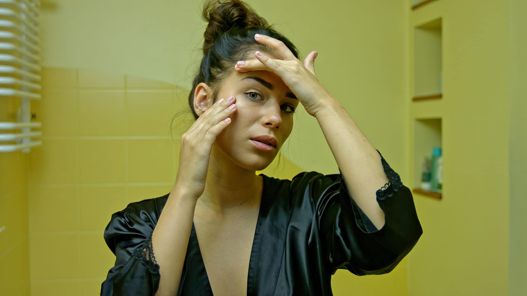 A young woman in the home bathroom looks in the mirror and checks her first wrinkles