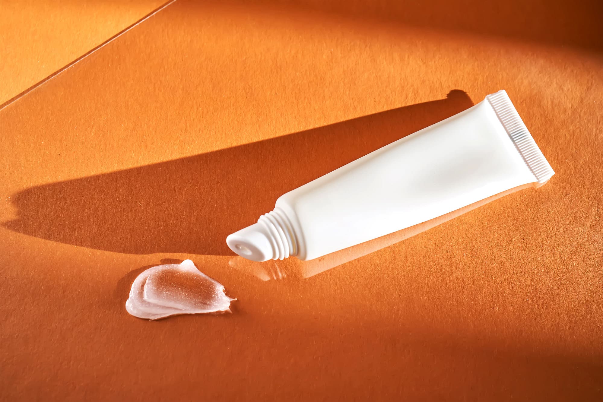 A white squeeze tube of acne cream rests on an orange surface, its shadows softly cast alongside the delicate swirl of cream