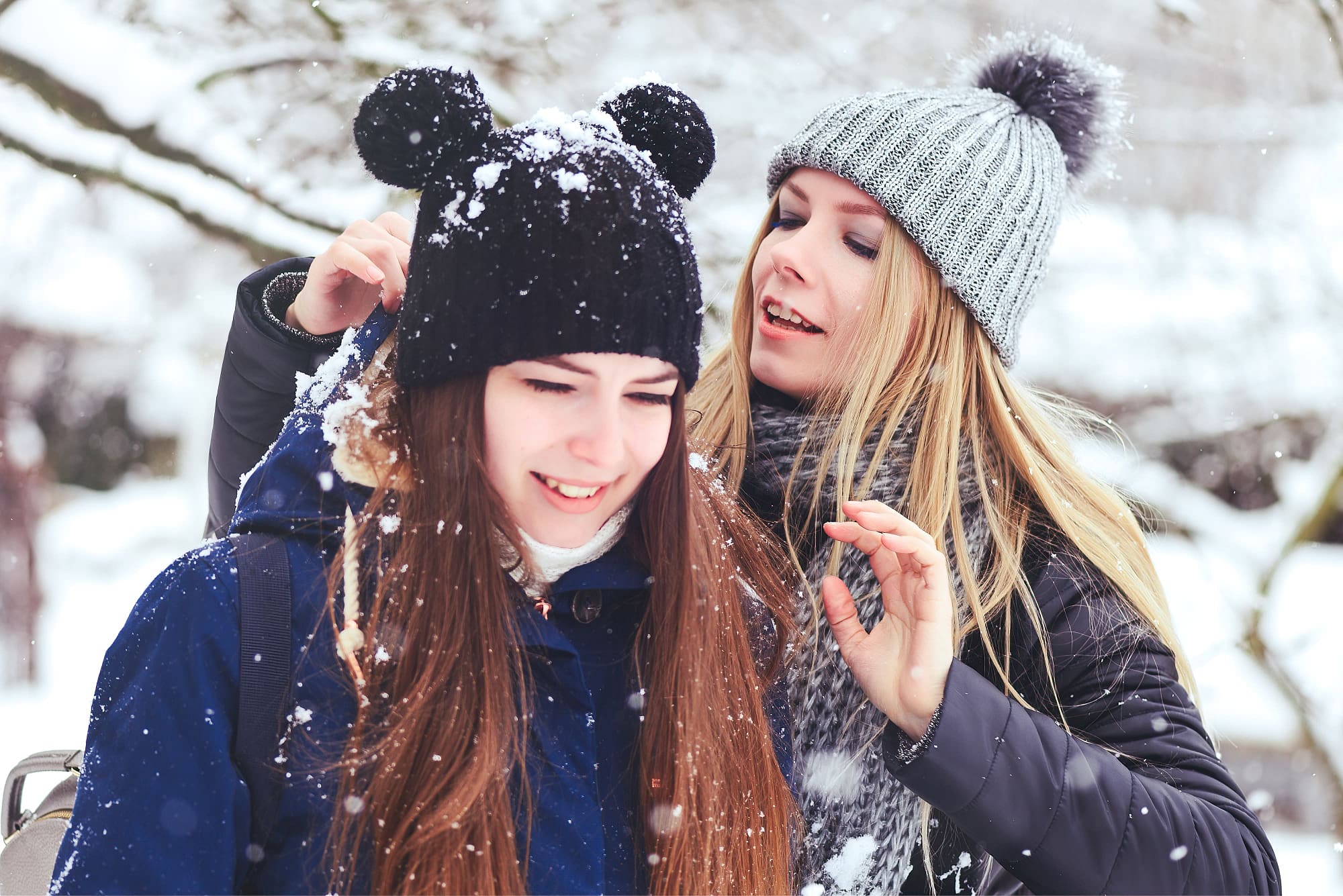 A woman and a girl in winter attire, with the woman brushing snow off the girl's beanie and jacket, standing outdoors in a snowy setting