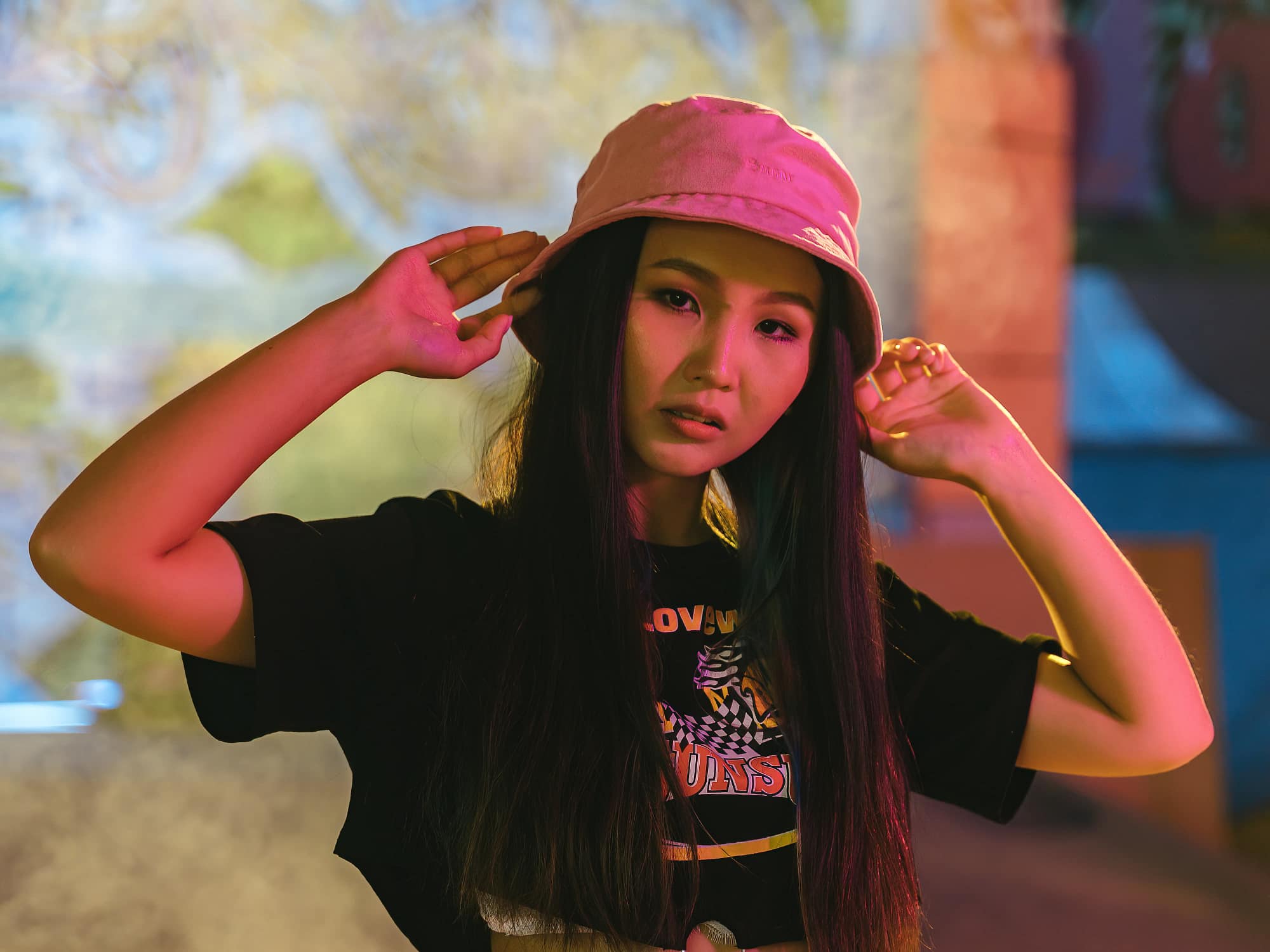 A woman with long dark hair sports a stylish pink bucket hat and a black T-shirt, posing with her hands near her head. The background features colorful, blurred lights