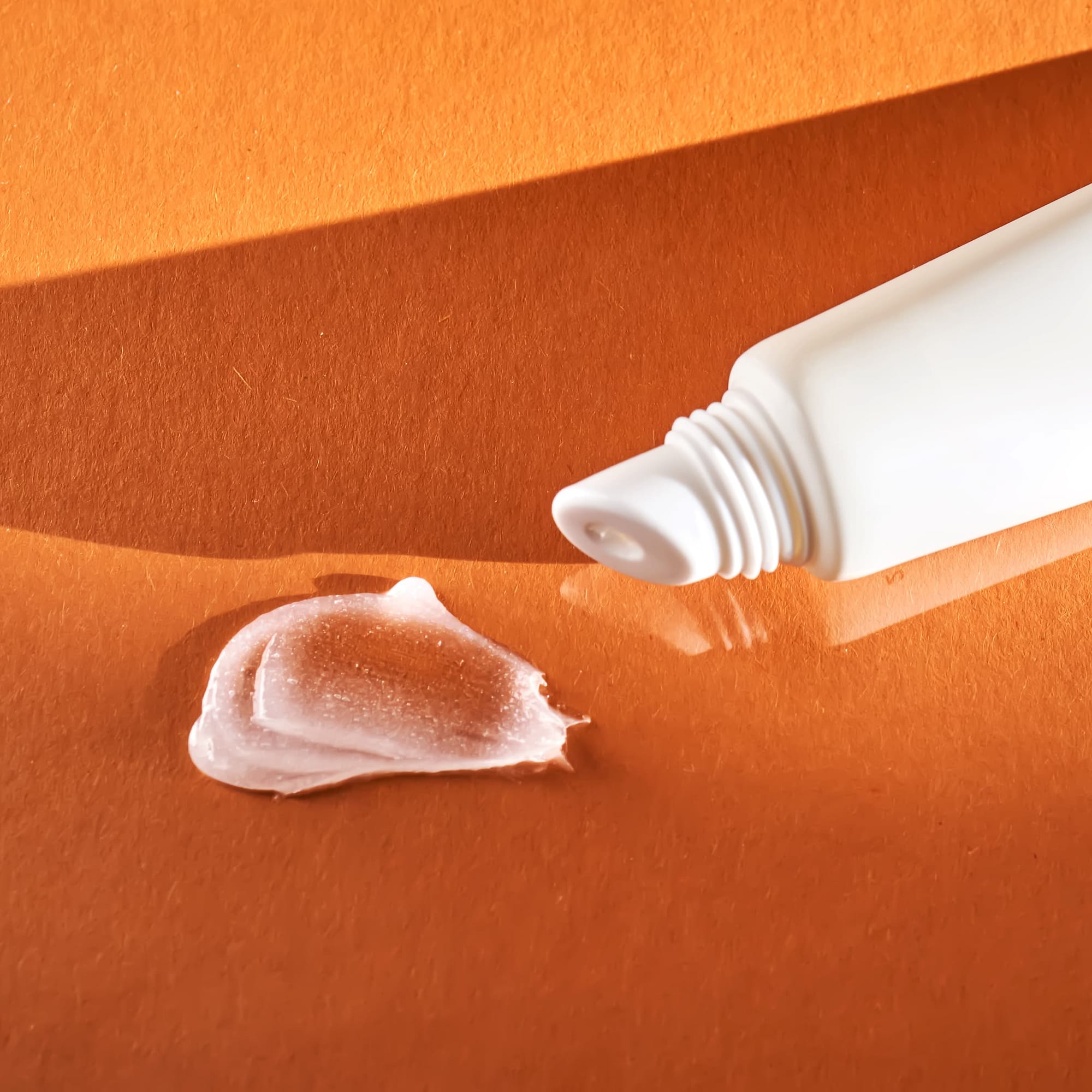 A white squeeze tube of acne cream rests on an orange surface, its shadows softly cast alongside the delicate swirl of cream