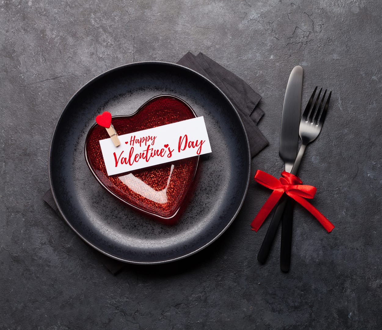 Heart-shaped dessert on a black plate with a "Happy Valentine's Day" card. Black napkin and cutlery tied with red ribbon beside the plate on a dark textured surface