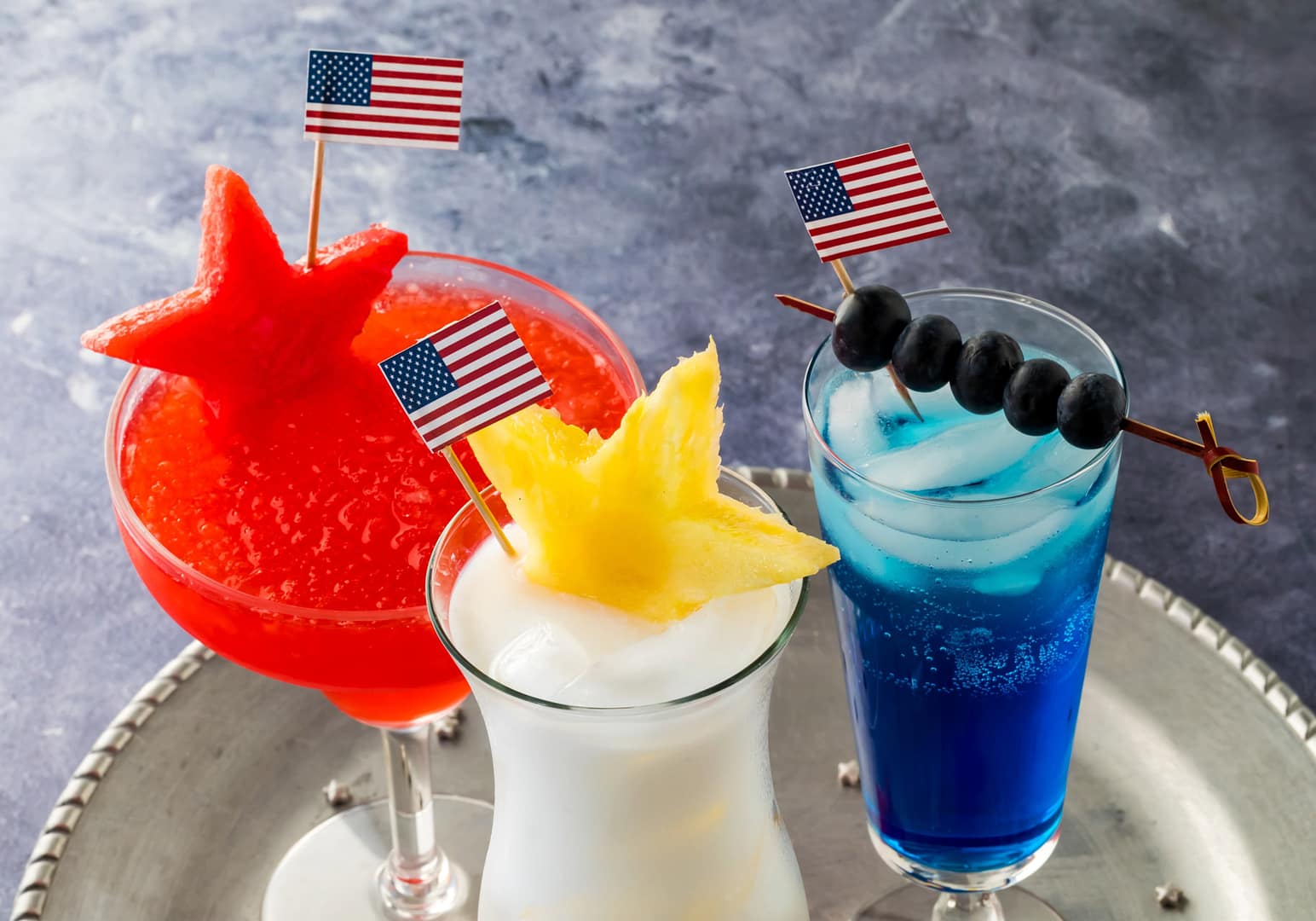 Three Fourth of July cocktails garnished with USA flags and fresh fruit