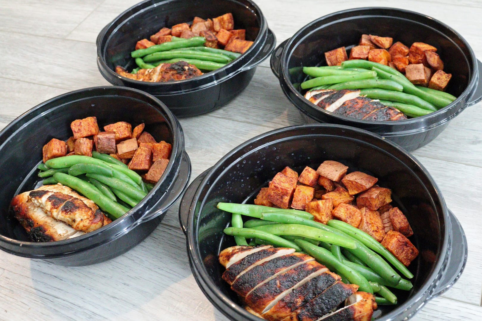 Bodybuilding meal prep dishes in four black bowls on a light gray wooden dining table