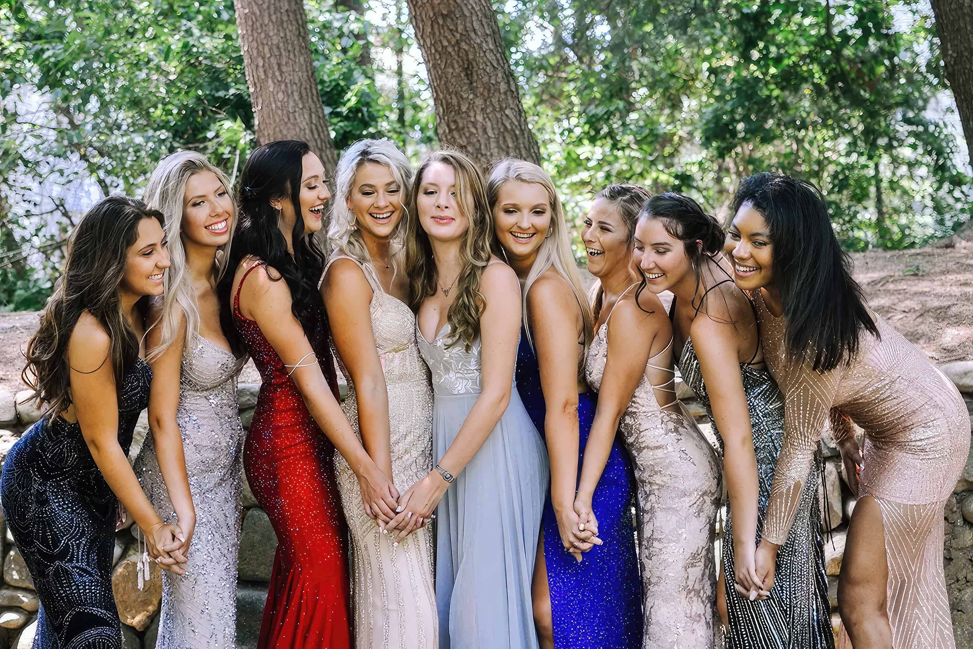 Nine teenage girls in elegant prom dresses stand in a line, holding hands and smiling, in an outdoor, wooded area