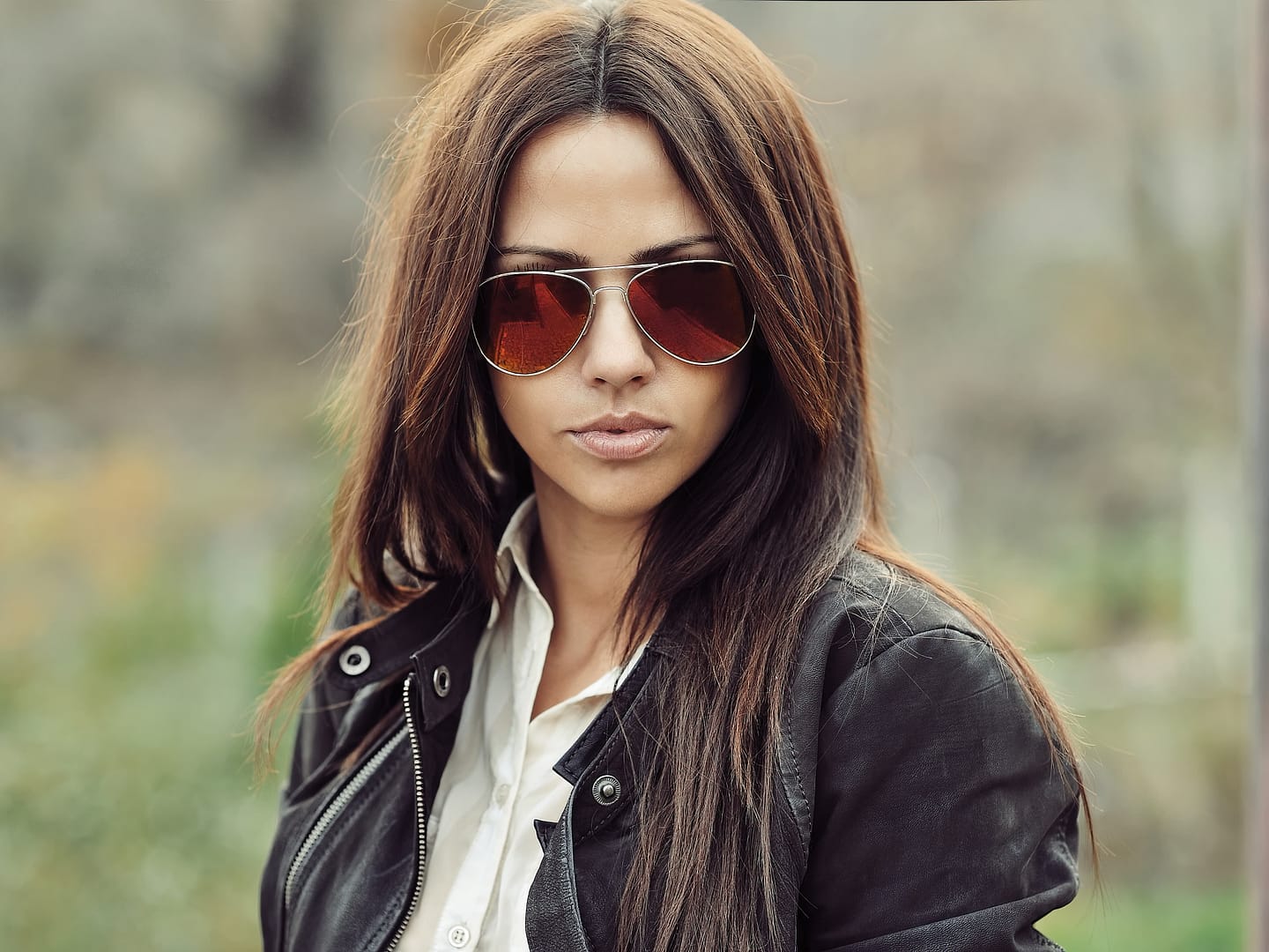 Woman with long brown hair wearing aviator sunglasses and a black jacket, standing outdoors