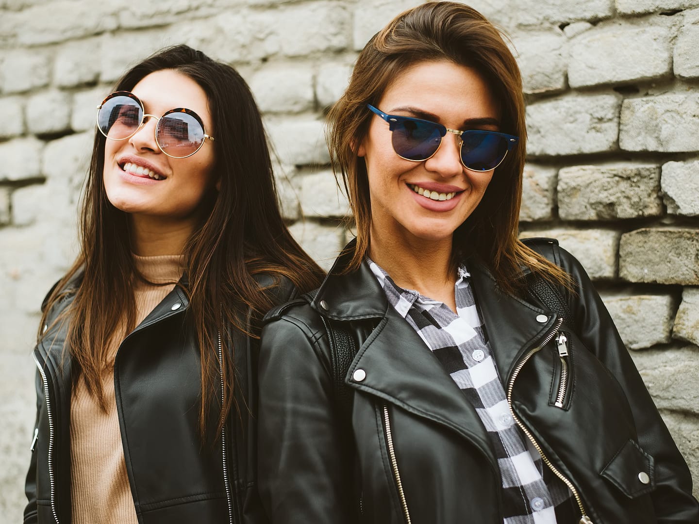 Two women wearing sunglasses and black leather jackets smile while standing in front of a light grey brick wall