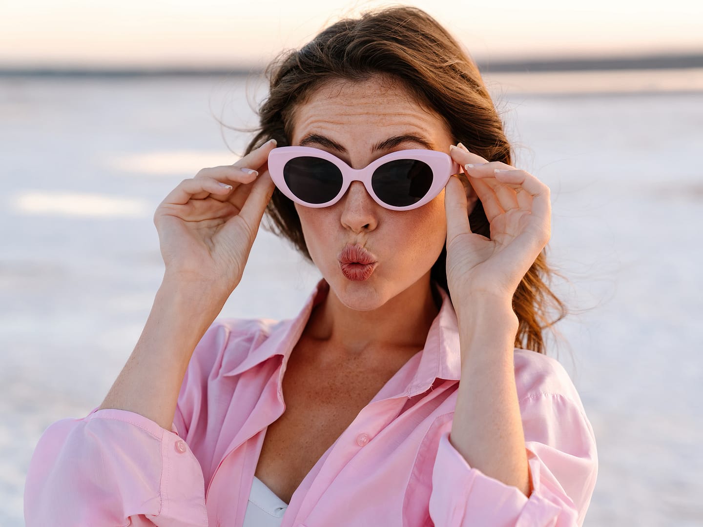 Woman wearing pink oval sunglasses and a pink shirt puckers her lips, standing outdoors with a blurred background