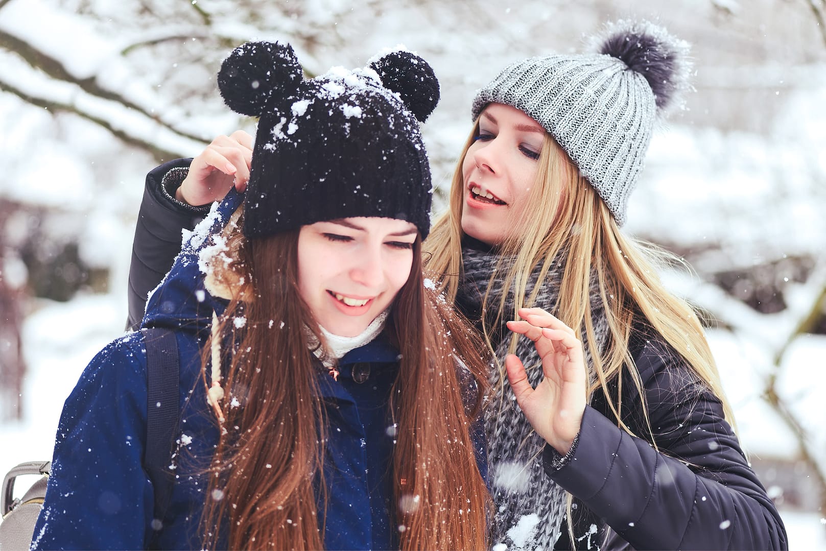 A woman and a girl in winter attire, with the woman brushing snow off the girl's beanie and jacket, standing outdoors in a snowy setting