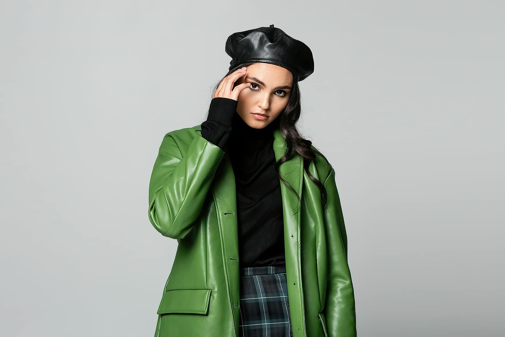 Woman wearing a black beret, green leather coat, black top, and checkered pants stands against a plain background, embodying the essence of fall fashion