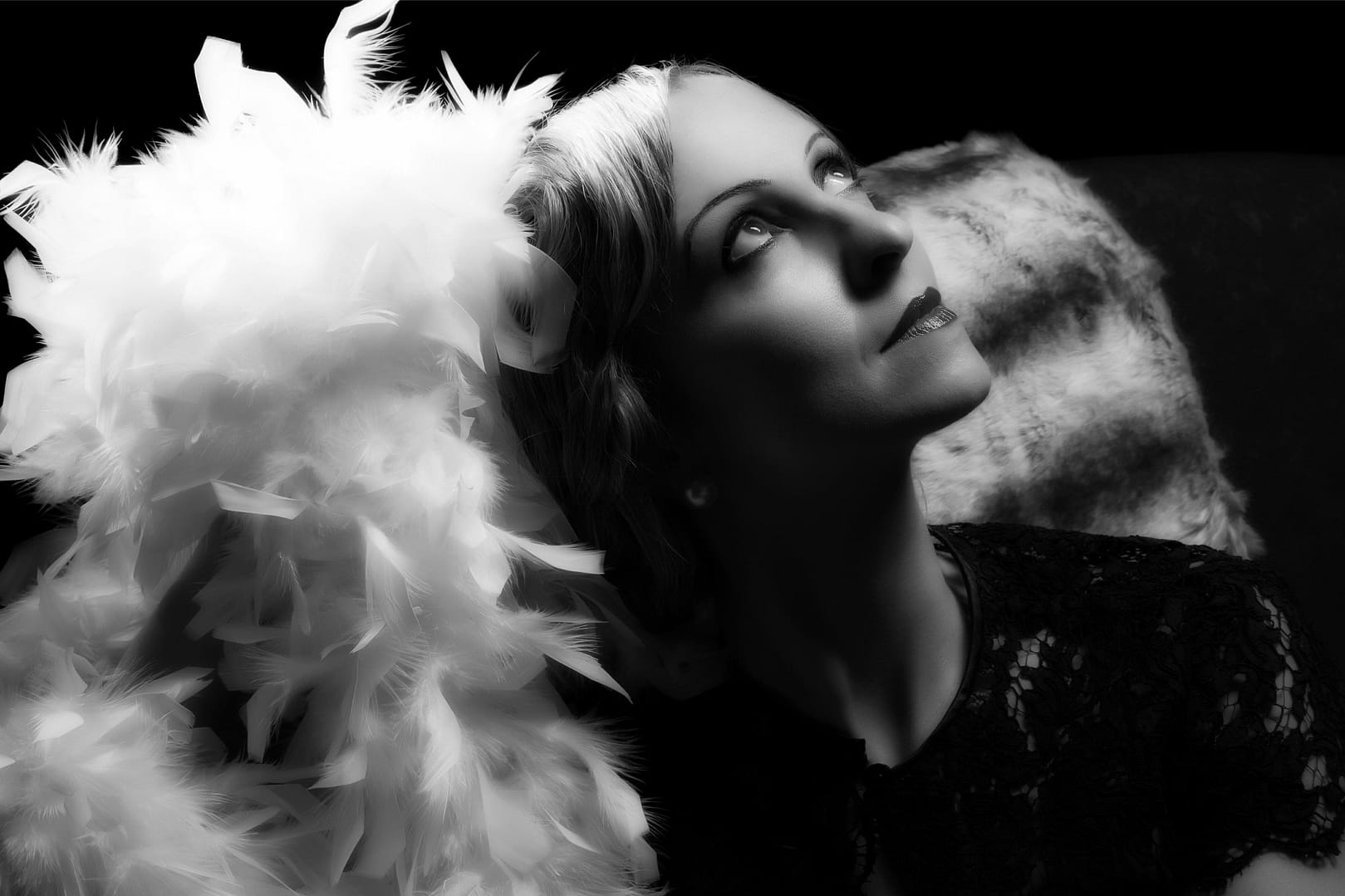 A classy woman in black attire looks up thoughtfully, holding a white feather boa in an elegant setting. The background is dark, accentuating her contemplative expression and the boa's texture
