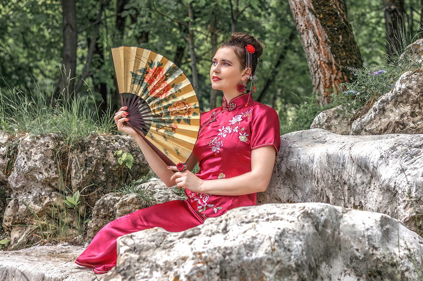 A woman in a red traditional Cheogsam dress sits on rocky terrain, holding an ornate folding fan. She has dark hair adorned with red flowers, and there is forest greenery in the background