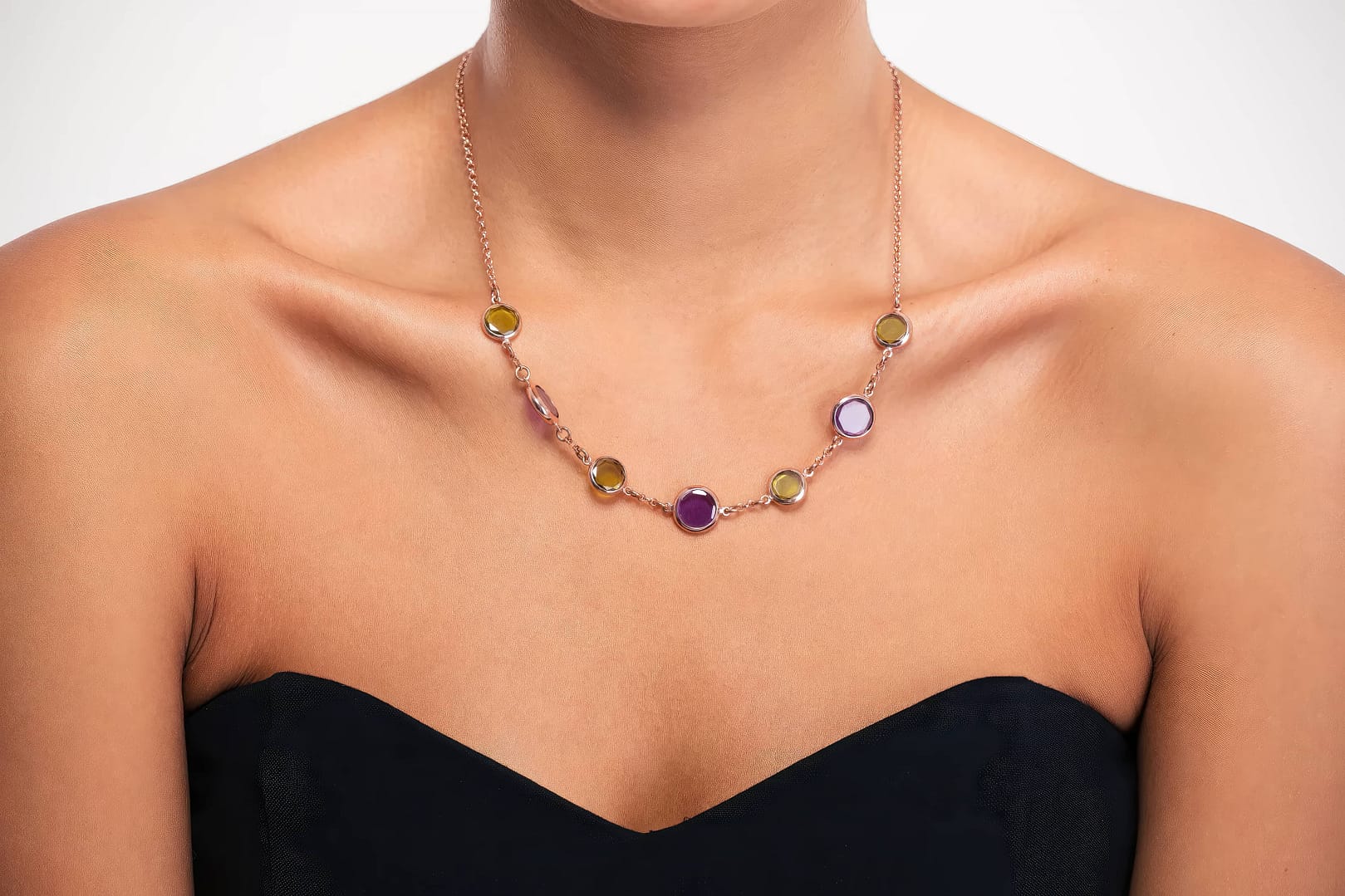 A woman wearing a strapless black top showcases a necklace with multicolored gem stones set on a gold chain against a plain background