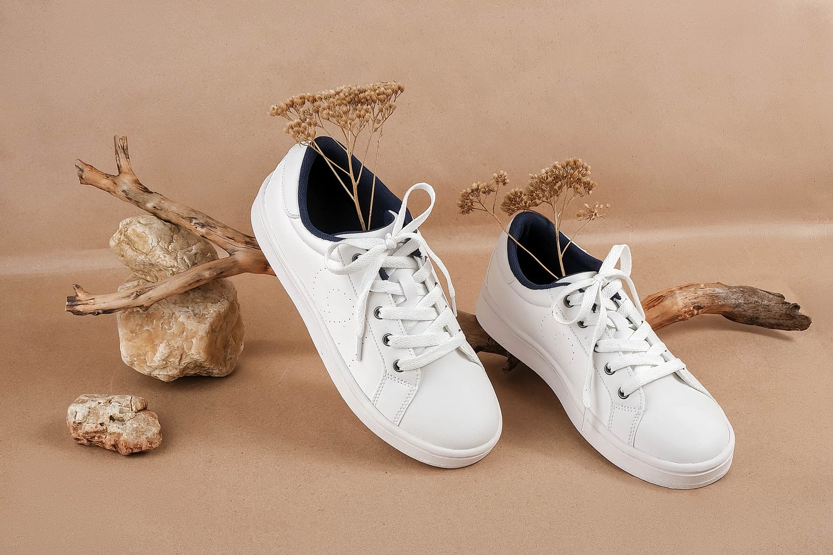 A pair of white sneakers with dry flowers on the wood and pile of stones, neutral beige craft paper background