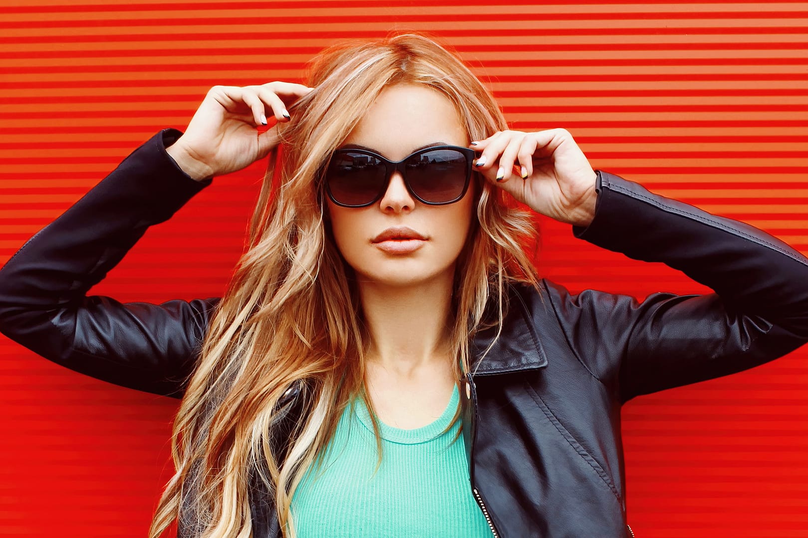 Stylish portrait of beautiful blonde woman wearing a black rock jacket and sunglasses on red background