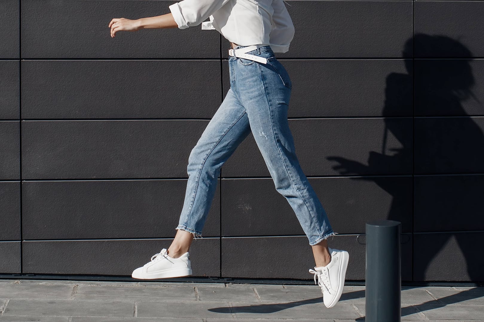 Woman dressed in a white shirt and mid-rise blue jeans is captured mid-stride while walking past a black tiled wall. The shadow of the woman is cast on the wall