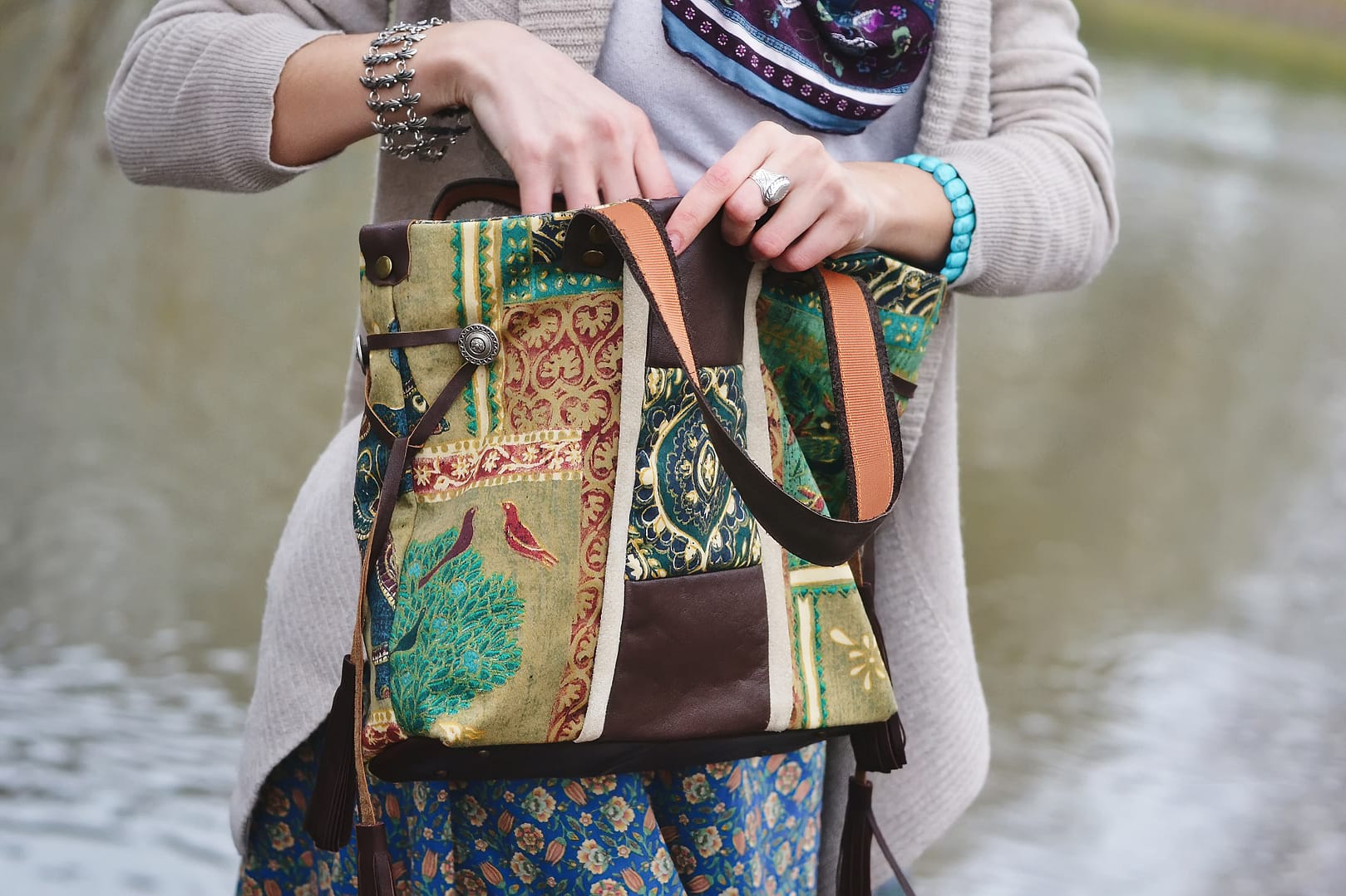 A person in a light cardigan and patterned skirt is holding a multi-colored, patchwork designer tote bag near a body of water