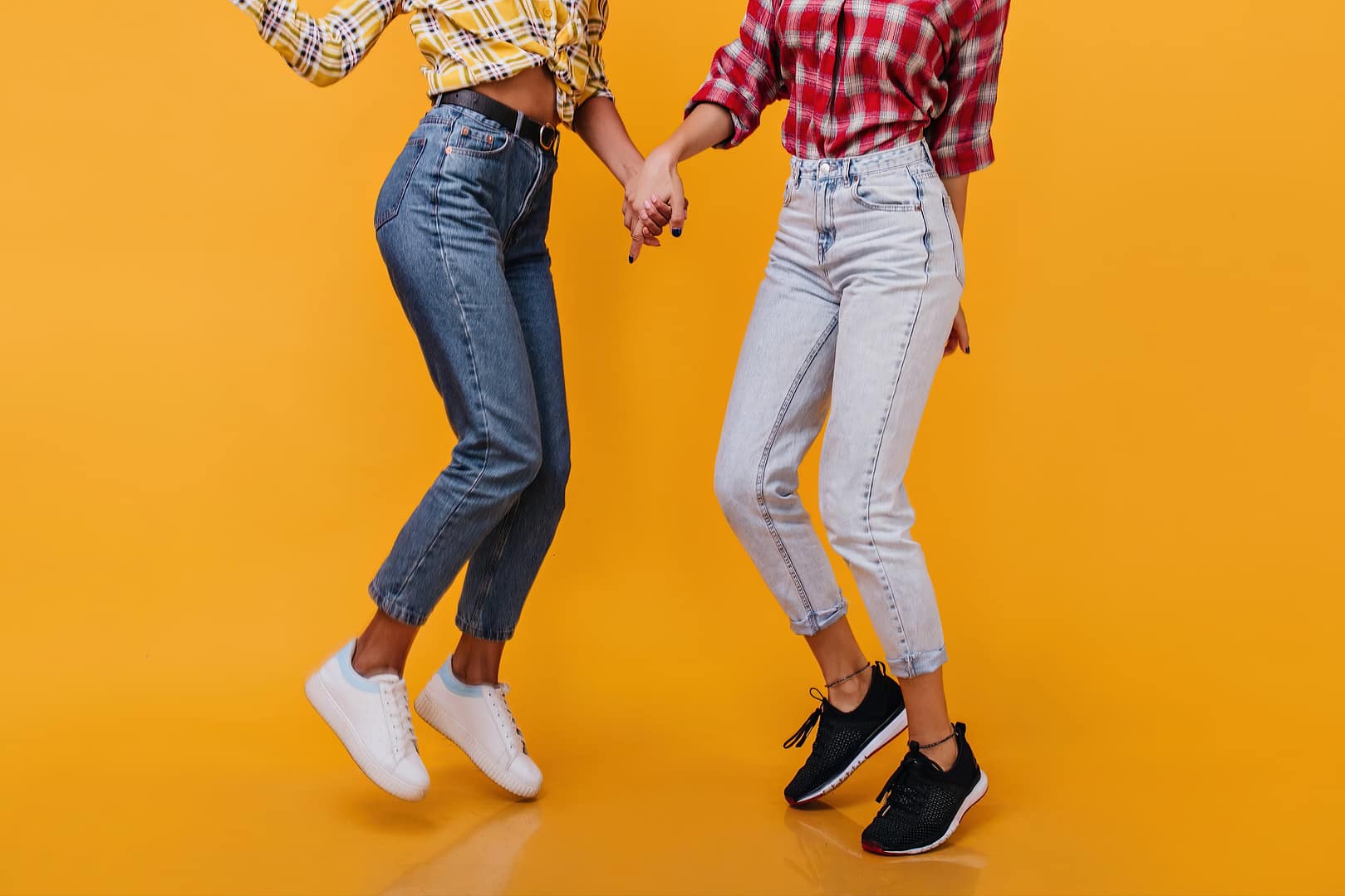 Two people holding hands are mid-jump against a bright orange background, wearing plaid shirts and high-waisted jeans. Their upper bodies are not shown