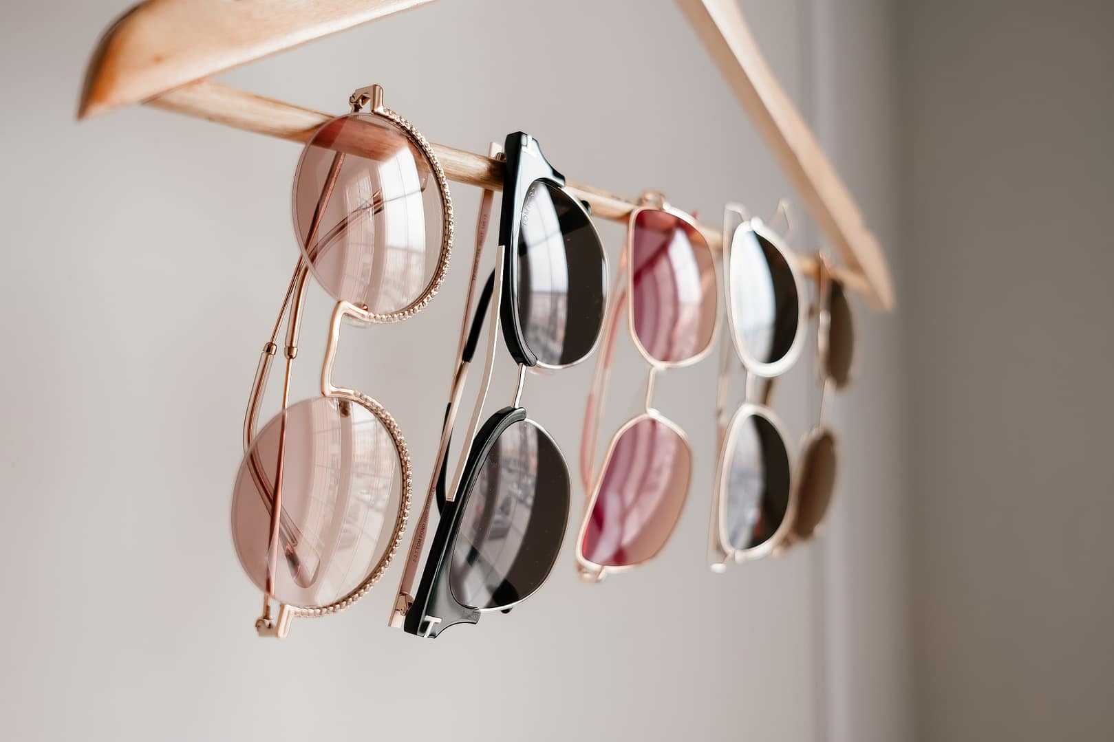 Four pairs of sunglasses in various styles hang from a wooden hanger against a plain background