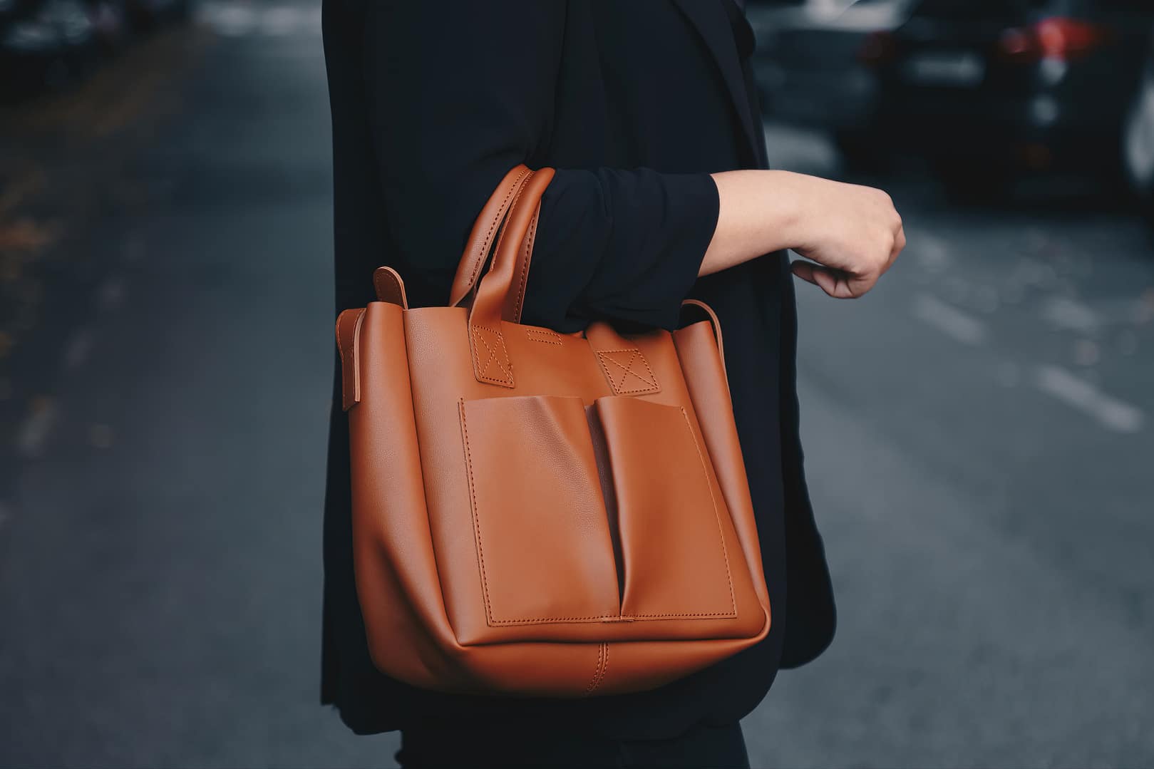 Close-up of a brown leather work bag from a woman in black outfit holding the bag walking on a street