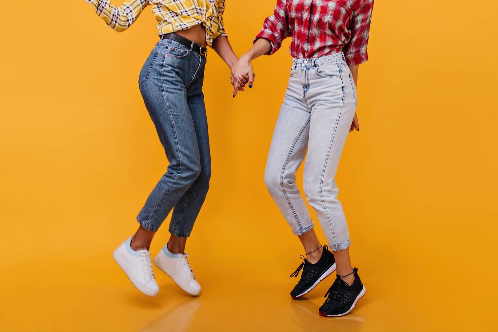 Two people holding hands are mid-jump against a bright orange background, wearing plaid shirts and high-waisted jeans. Their upper bodies are not shown