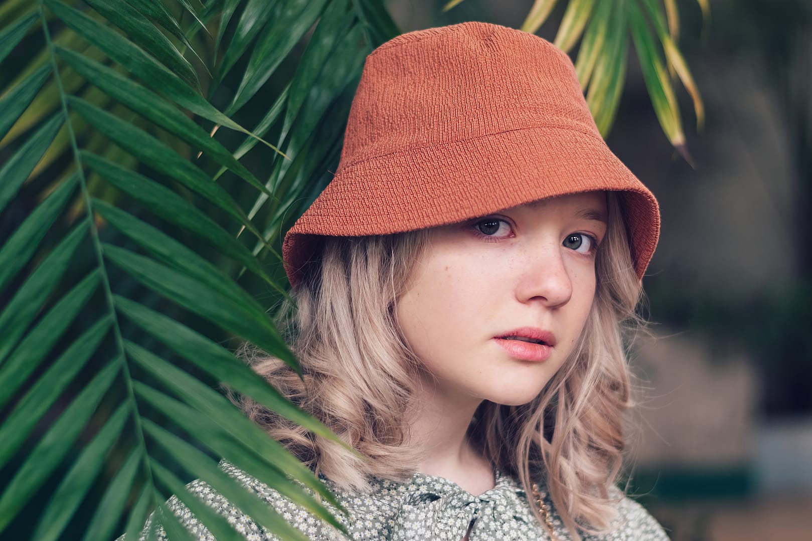 A young woman with blonde hair, wearing a bucket hat and a green patterned top, is partially obscured by large green leaves