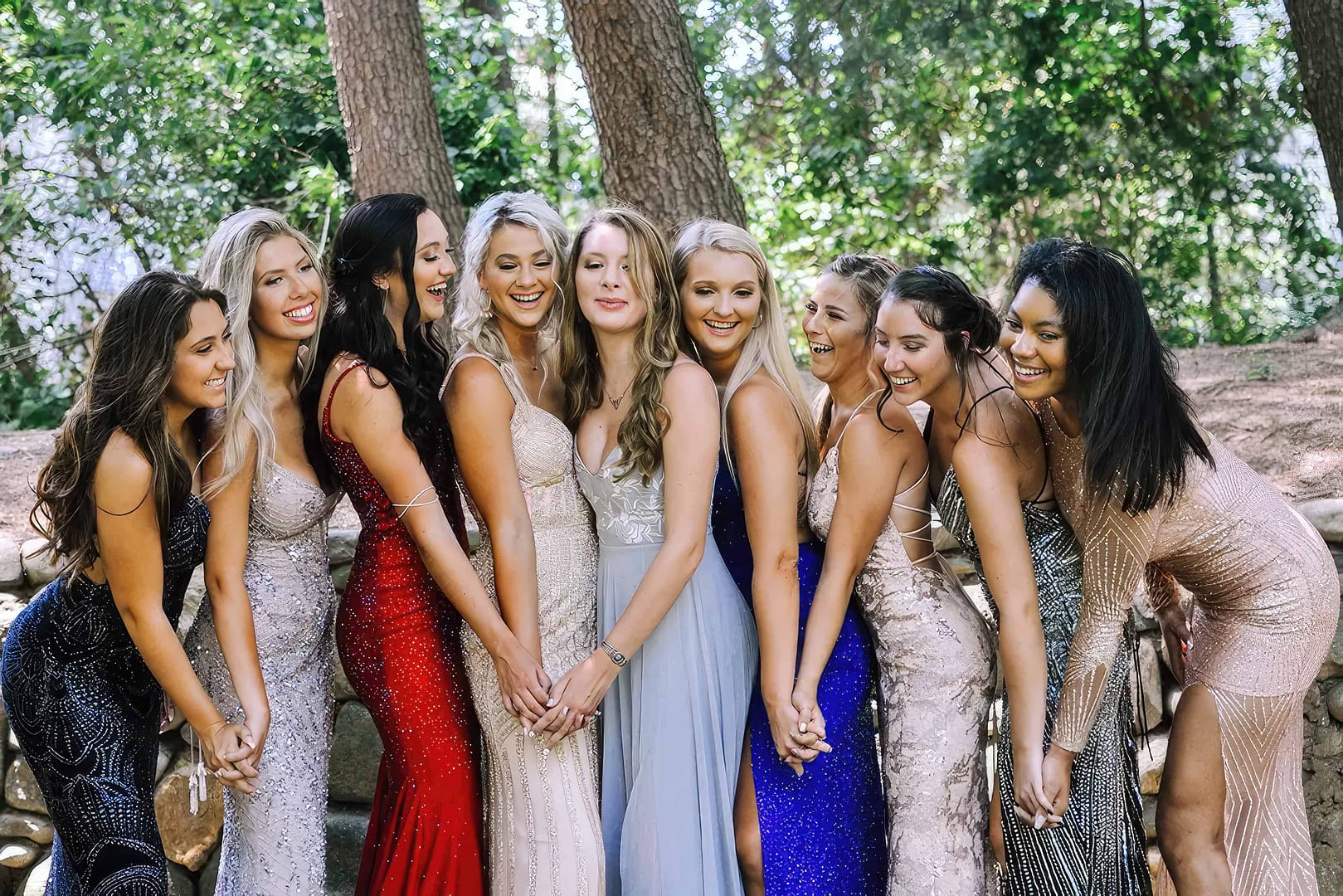 Nine teenage girls in elegant prom dresses stand in a line, holding hands and smiling, in an outdoor, wooded area