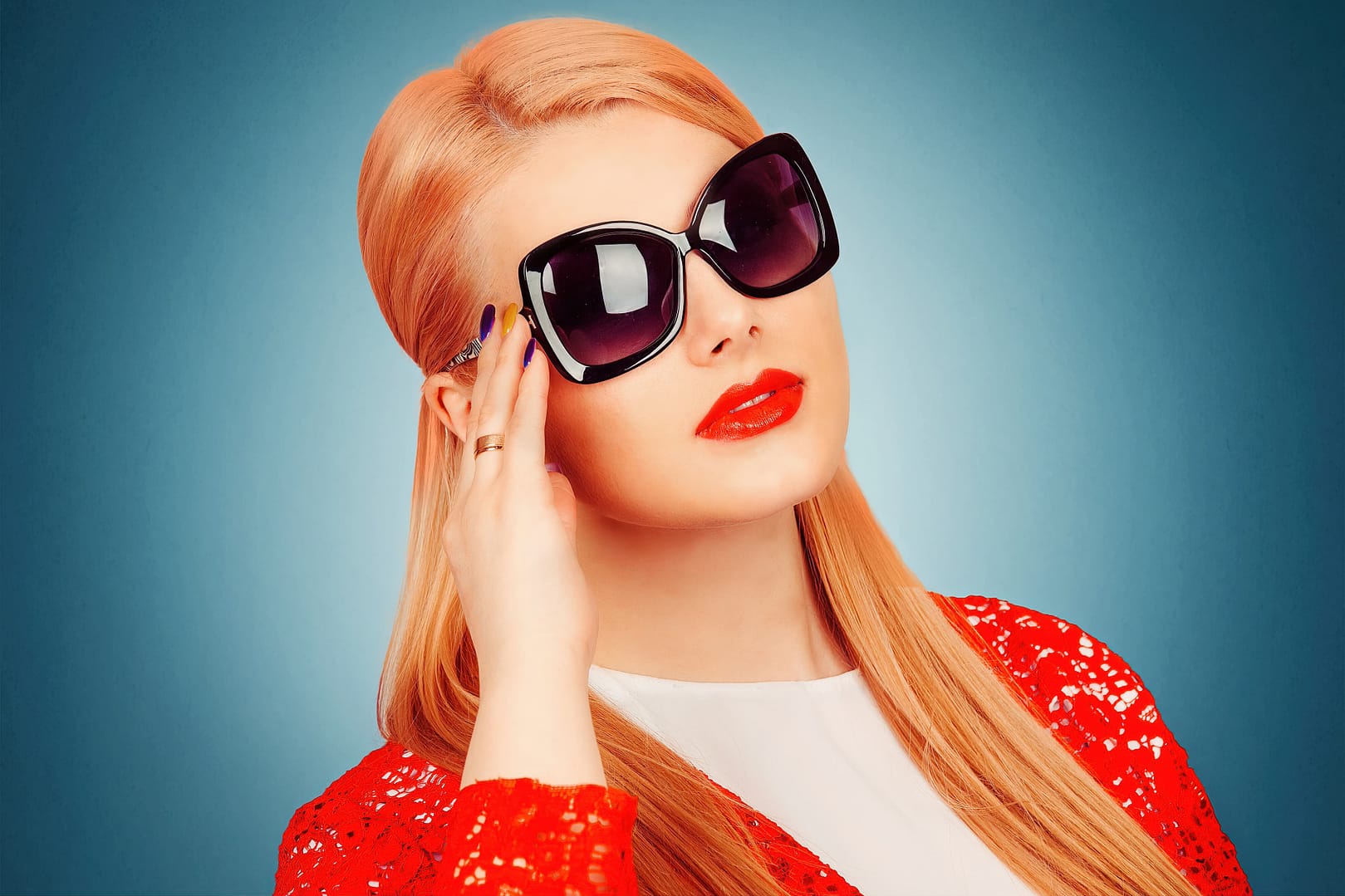 Woman with long blonde hair wearing large black sunglasses and a red jacket with a white top poses against a blue background